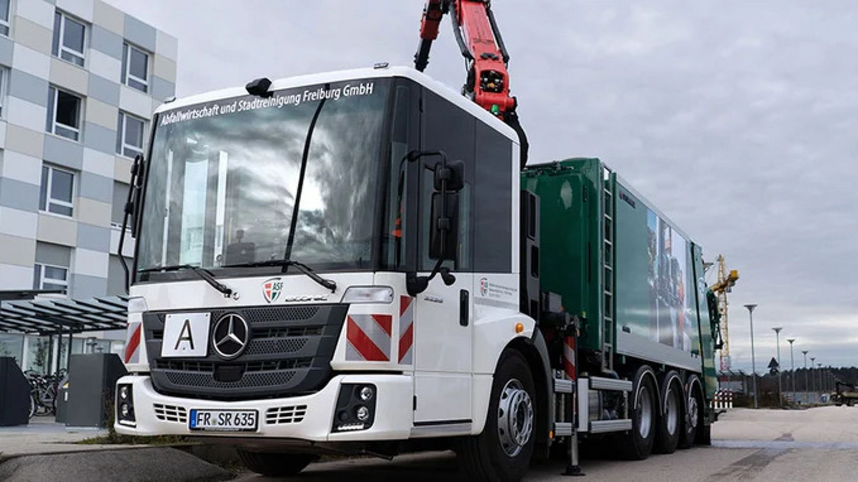 A Mercedes-Benz Econic refuse truck is parked in a parking lot. The truck has a green container with a raised crane arm.