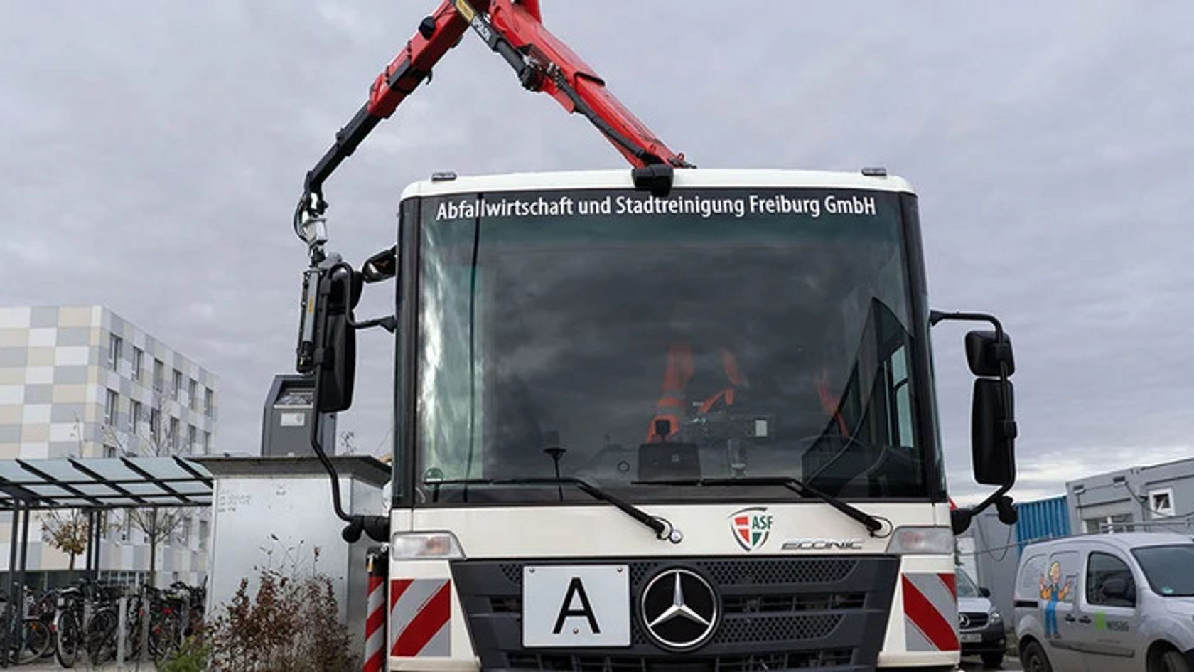 An Econic refuse collection vehicle of Abfallwirtschaft und Stadtreinigung Freiburg GmbH, photographed from the front. The truck vehicle has a crane arm on the left side that is used for lifting waste containers.