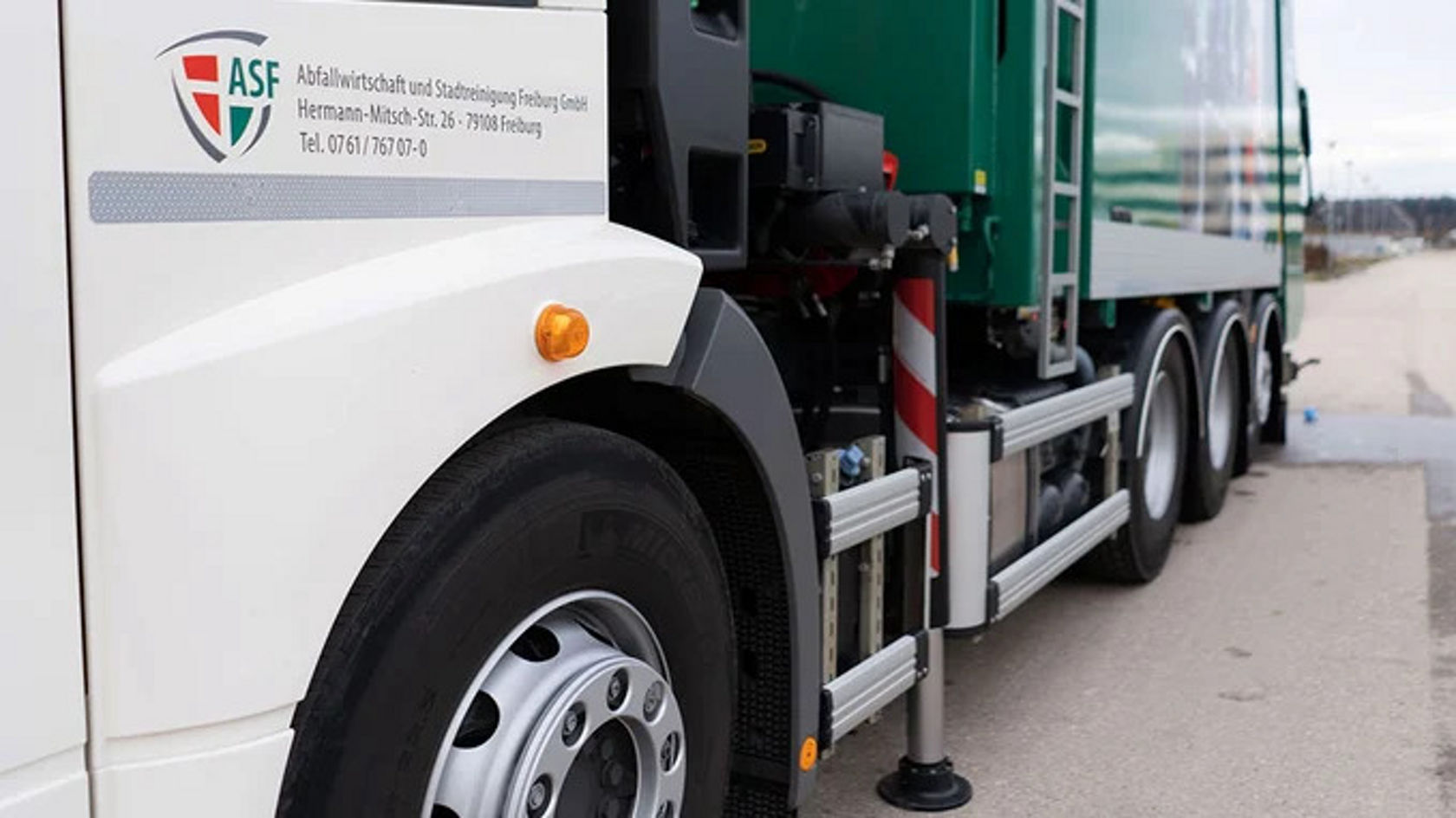 Close-up of a truck wheel and the side of an Econic refuse truck. The truck has a green loading area and is on a paved path.