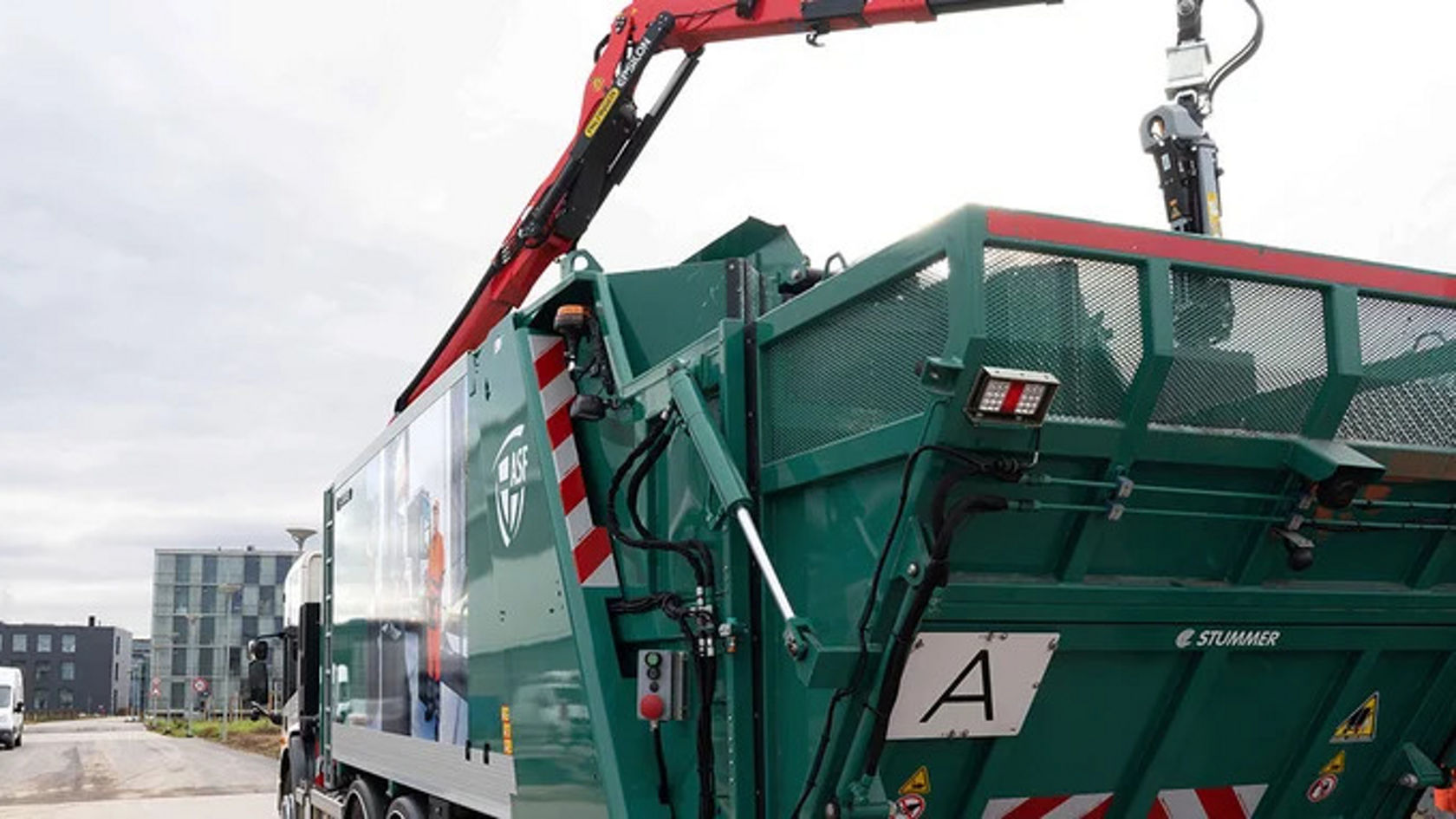 An Econic refuse truck with a crane arm stands in an urban environment. The truck has a green loading area with a large opening.