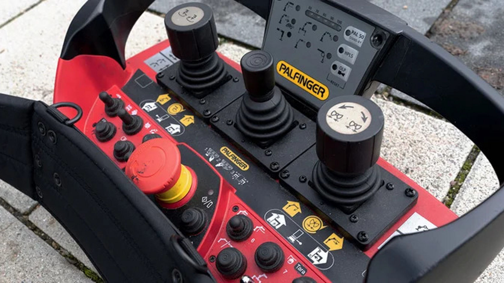 Remote control for a machine control system, with several joysticks and various red and black buttons, placed on a sidewalk.