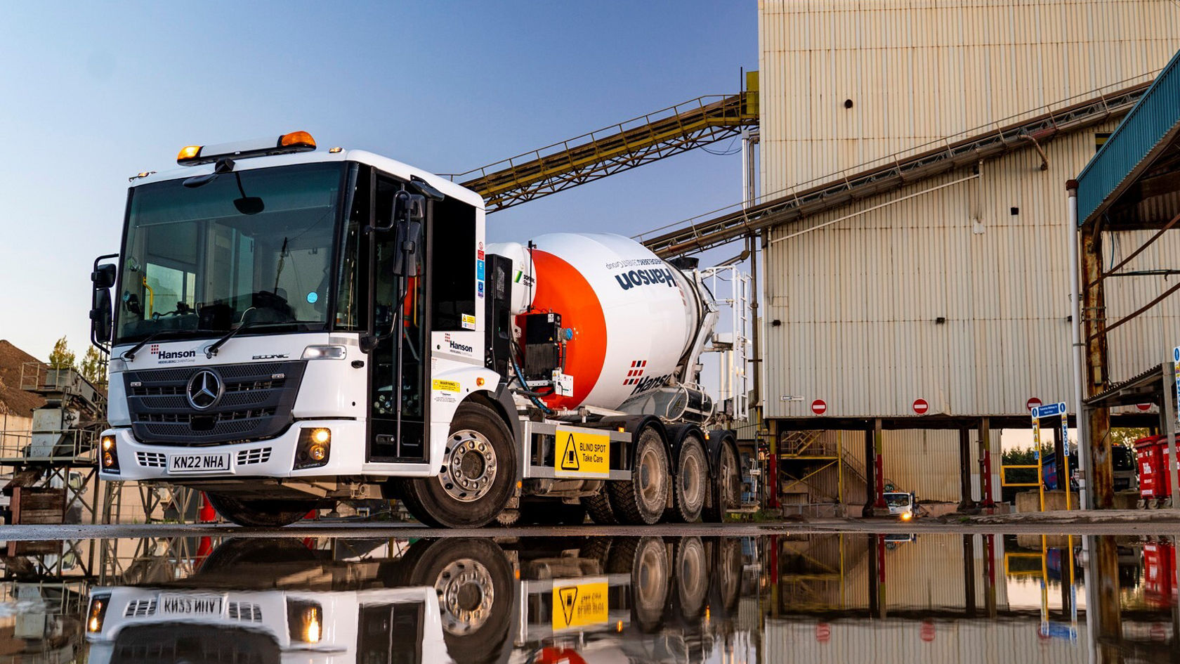 Ein Mercedes-Benz Econic Betonmischer steht an einem Industriegebäude, spiegelnd im Wasser. Der Lkw hat ein auffälliges Logo und Sicherheitswarnungen auf der Seite.