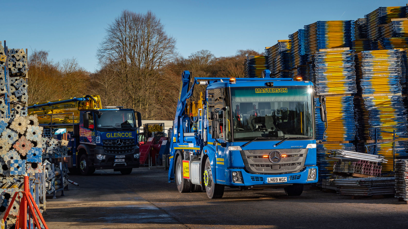 Zwei Lastwagen stehen auf einem Baustellenareal, einer mit dem Logo von Mercedes-Benz. Der blaue Lkw hat einen Kran, während der andere Lkw in Schwarz mit der Aufschrift "GLENCOE" fährt.