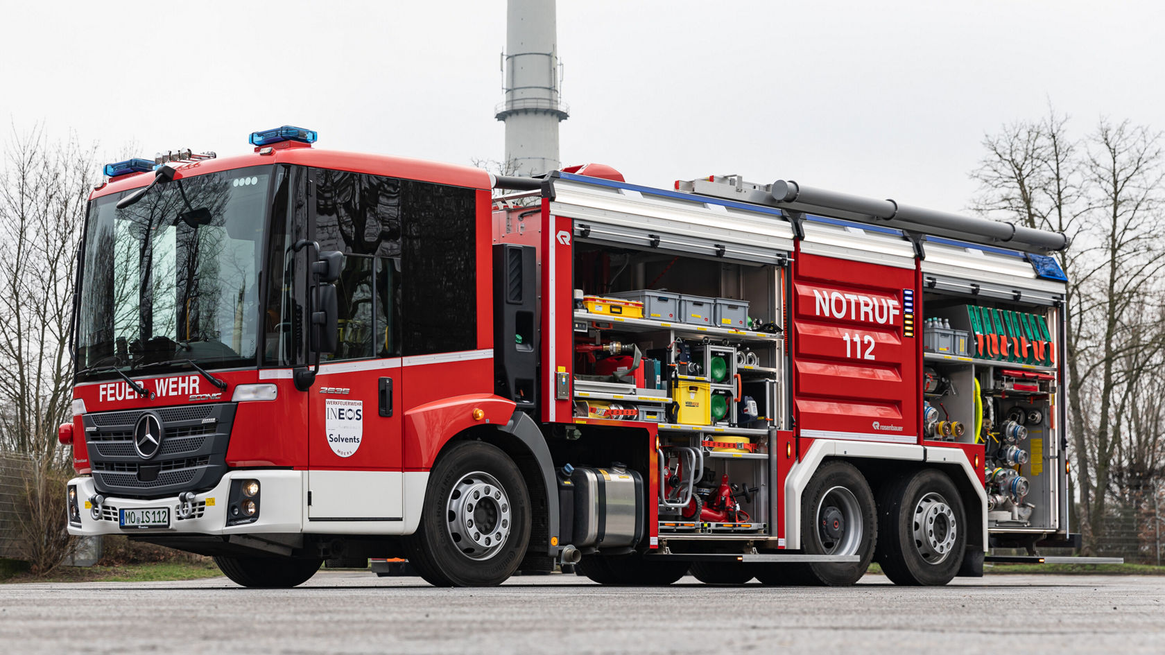 Véhicule de pompiers Mercedes‑Benz avec les portes latérales ouvertes, laissant apparaître le matériel de secours et de lutte contre l'incendie. Le véhicule est stationné sur une route, entouré d'arbres.
