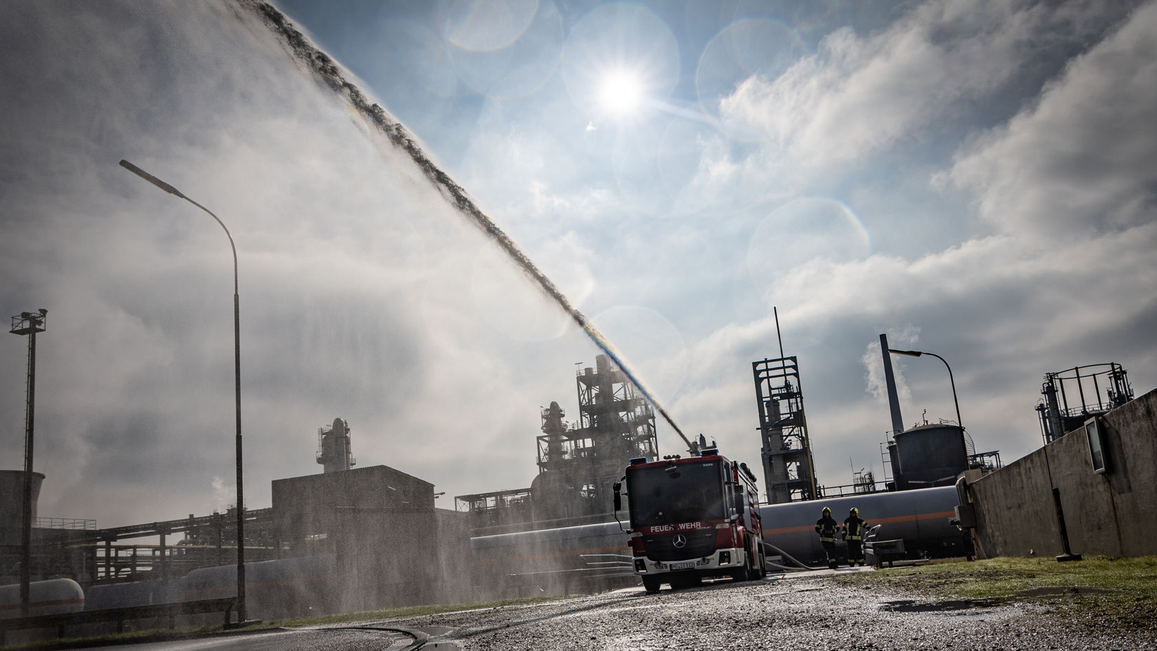 Un véhicule de pompiers projette de l'eau dans une installation industrielle, avec de hautes cheminées d'usine et des structures métalliques en arrière-plan. Des pompiers travaillent au premier plan.