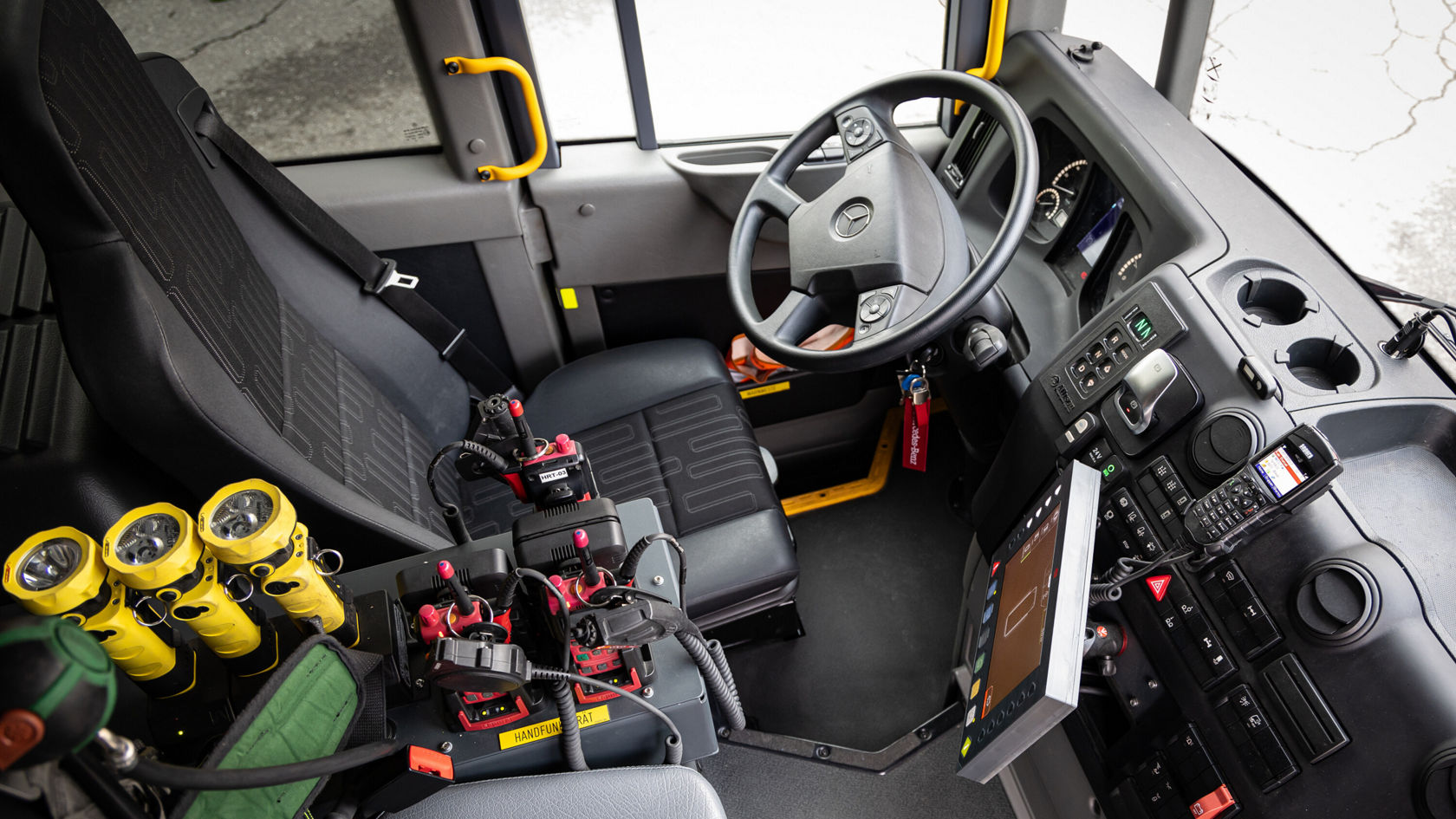 L'intérieur d'un véhicule Mercedes‑Benz montre un siège conducteur avec un volant, des éléments de commande et un afficheur. Sur le siège passager, plusieurs lampes jaunes et un équipement sont visibles.