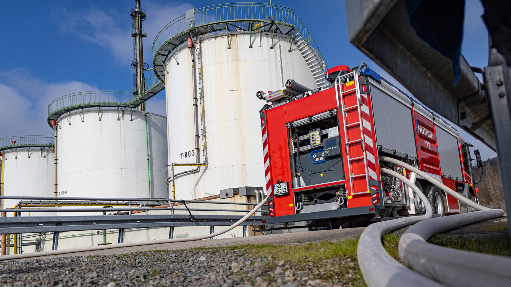 Un véhicule de pompiers est stationné près d'installations de stockage, des tuyaux y sont raccordés. De grands réservoirs de stockage blancs sont visibles en arrière-plan. La scène suggère une intervention dans le domaine de la lutte contre l'incendie ou de l'aide d'urgence.