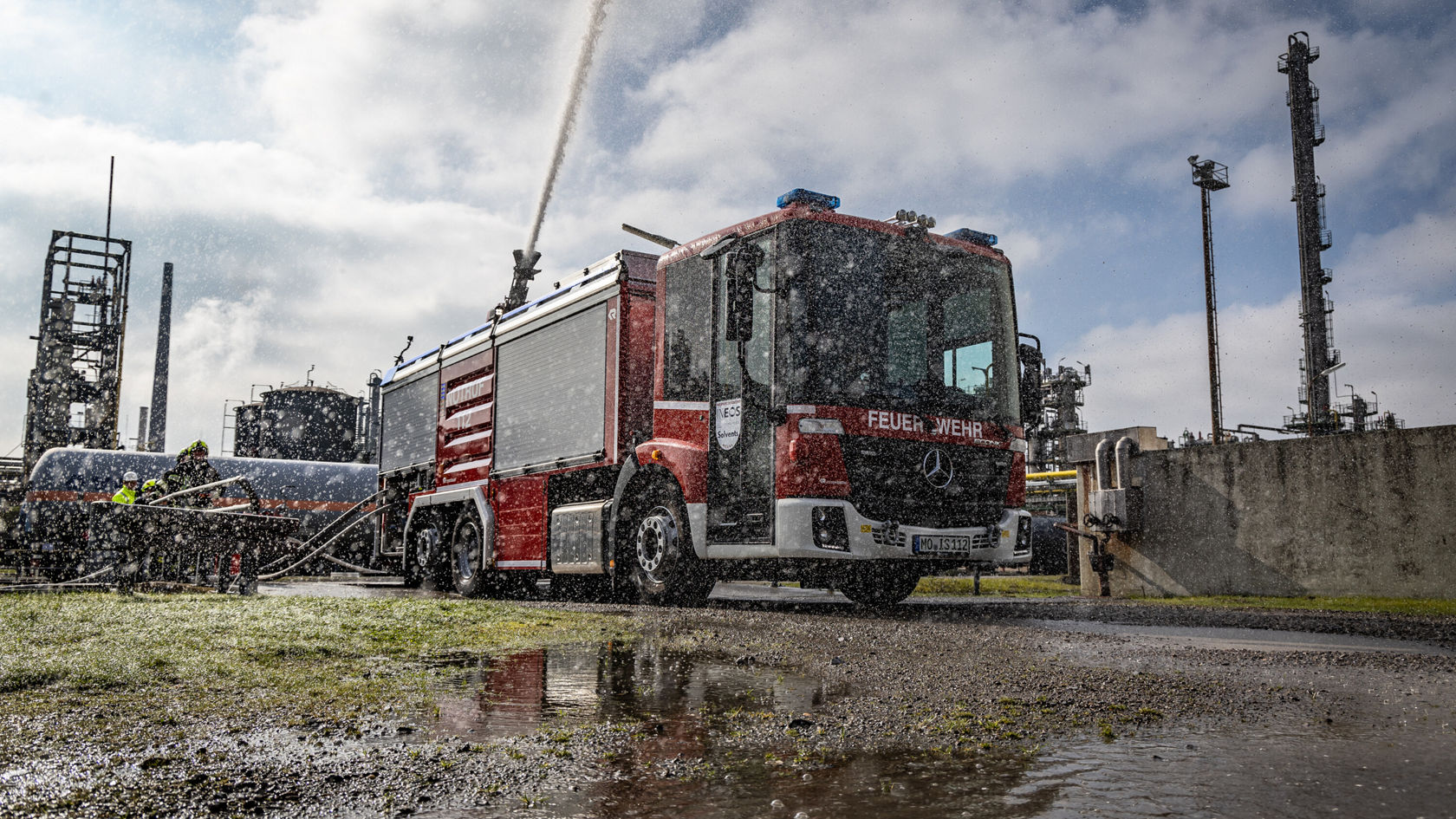 Un véhicule d'incendie Mercedes‑Benz rouge projette de l'eau sur un site d'intervention dans une installation industrielle, tandis que des pompiers travaillent à côté.