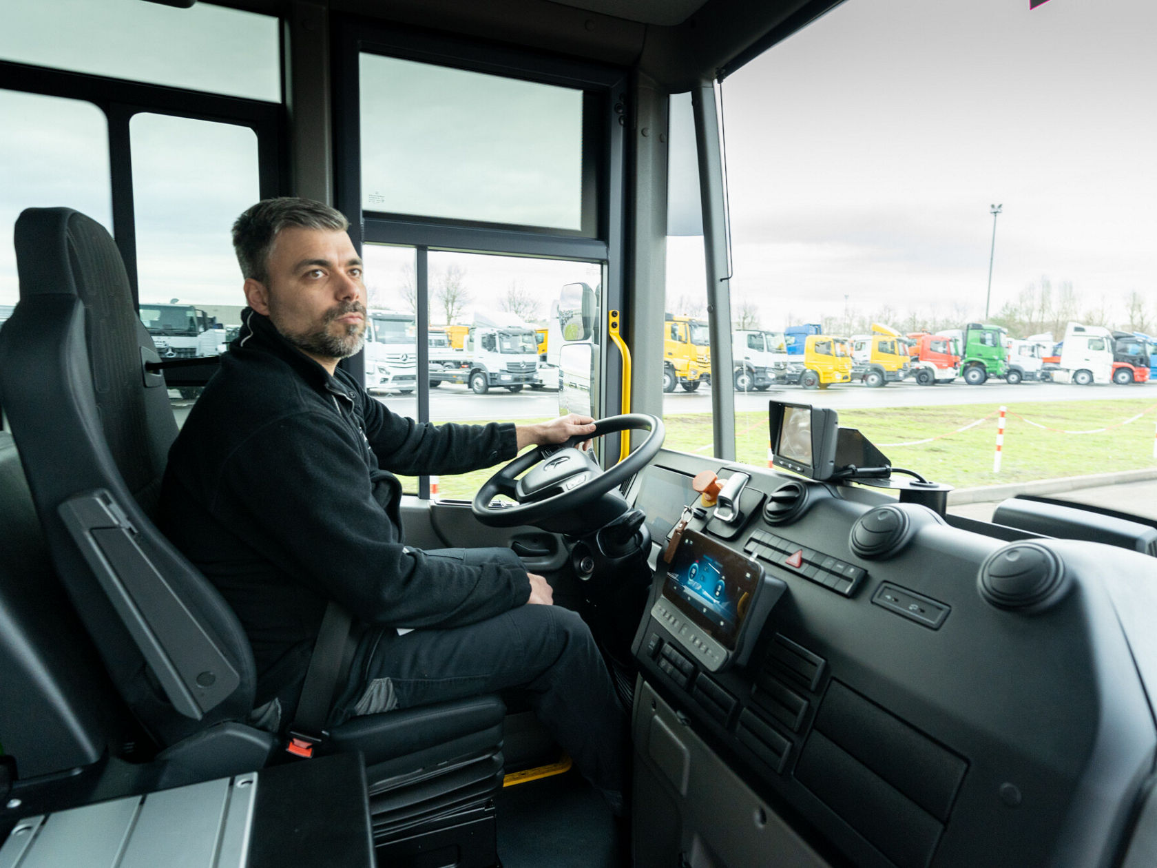 Ein Mann sitzt im Fahrersitz eines Mercedes-Benz Econic Trucks und blickt konzentriert nach vorne. Im Hintergrund sind weitere Lkw geparkt.
