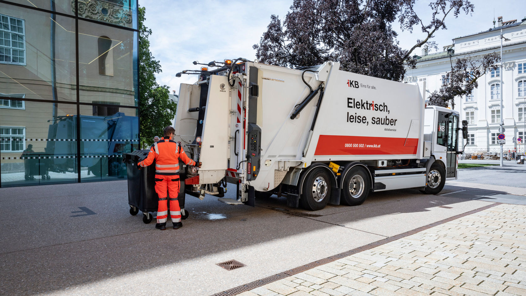 Ein eEconic Müllwagen steht auf einer Straße. Ein Mitarbeiter in orangefarbenem Arbeitsanzug entleert eine schwarze Mülltonne in den Laderaum des Fahrzeugs.