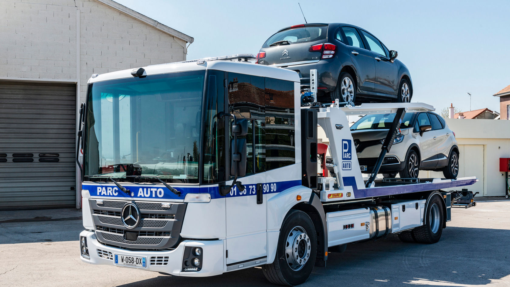Un camión Mercedes-Benz Econic transporta dos coches sobre una plataforma de carga mientras está estacionado en un aparcamiento para mostrar un servicio de transporte de vehículos.