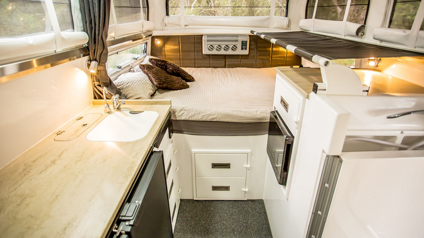 Innenansicht eines Wohnmobils mit einem Bett, das mit Kissen dekoriert ist. Es gibt eine Küche mit einem Waschbecken und einem Kühlschrank. Große Fenster sorgen für viel Licht.