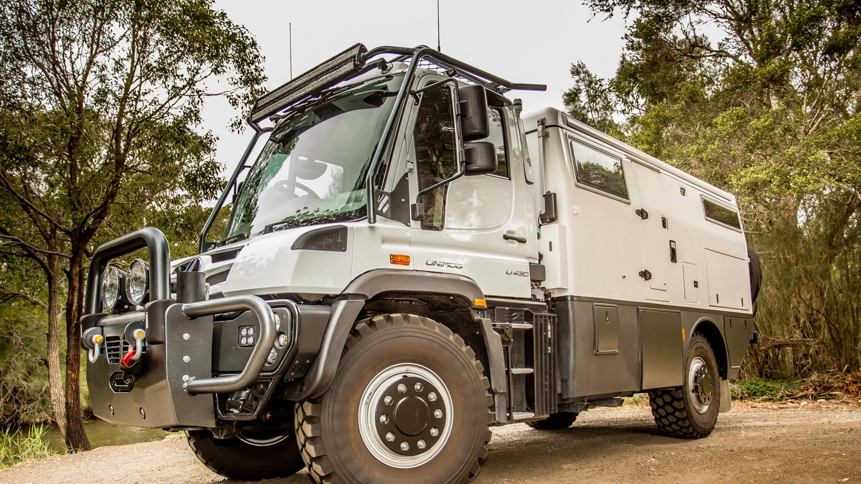 Ein Unimog U 430 Geländewagen mit Wohnmobil-Aufbau steht auf einem Schotterweg, umgeben von Bäumen und Natur.