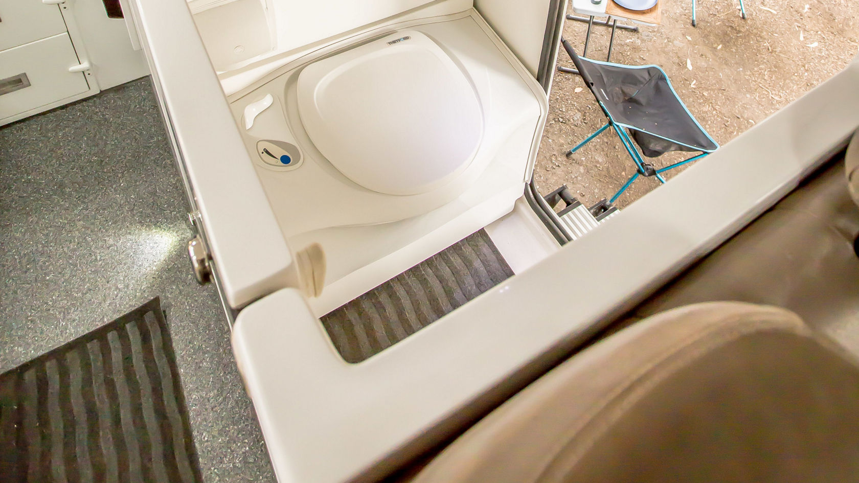 Bild zeigt das Innere eines Wohnmobils mit einem kompakten Badezimmer. Im Vordergrund befindet sich eine weiße Toilette, neben der eine Dusche mit Schlauch installiert ist. Im Hintergrund sind zwei Klappliegen und Geschirr auf einem Tisch im Freien sichtbar.