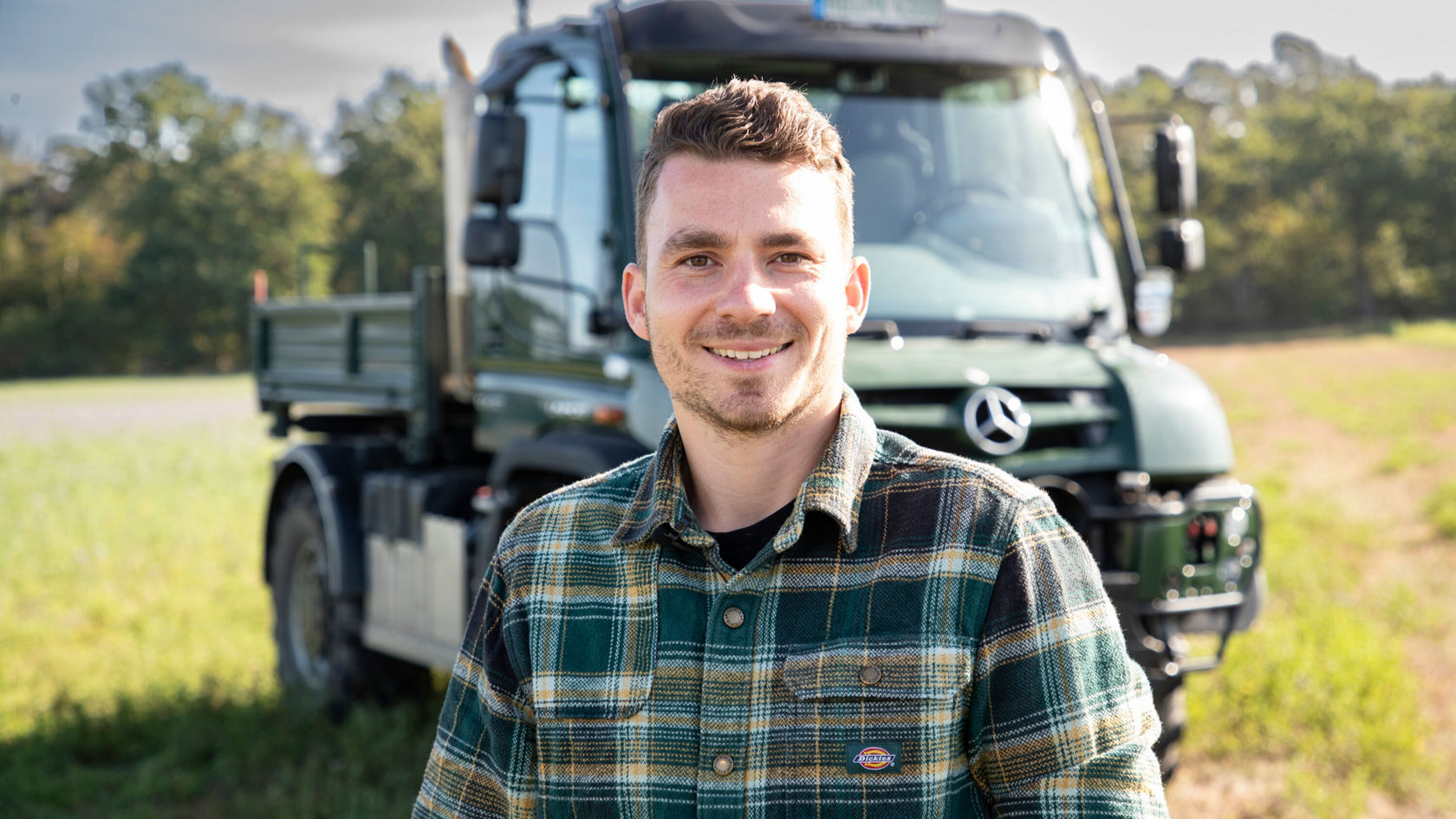 Ein lächelnder Mann steht auf einem Feld vor einem Mercedes-Benz Lkw, umgeben von Bäumen und natürlicher Landschaft. Der Mann trägt ein kariertes Hemd und wirkt freundlich.