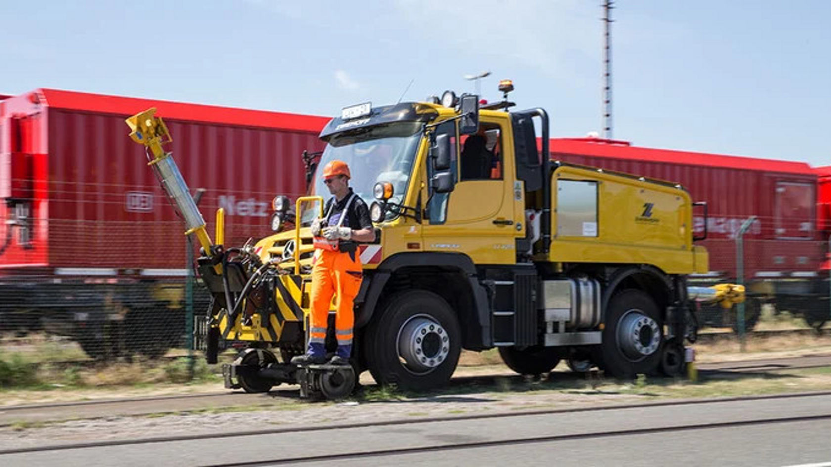 Ein Mercedes-Benz Unimog steht auf Gleisen, während ein Arbeiter in orangefarbener Kleidung einen Schwenkarm bedient. Im Hintergrund sind rote Bahncontainer zu sehen.