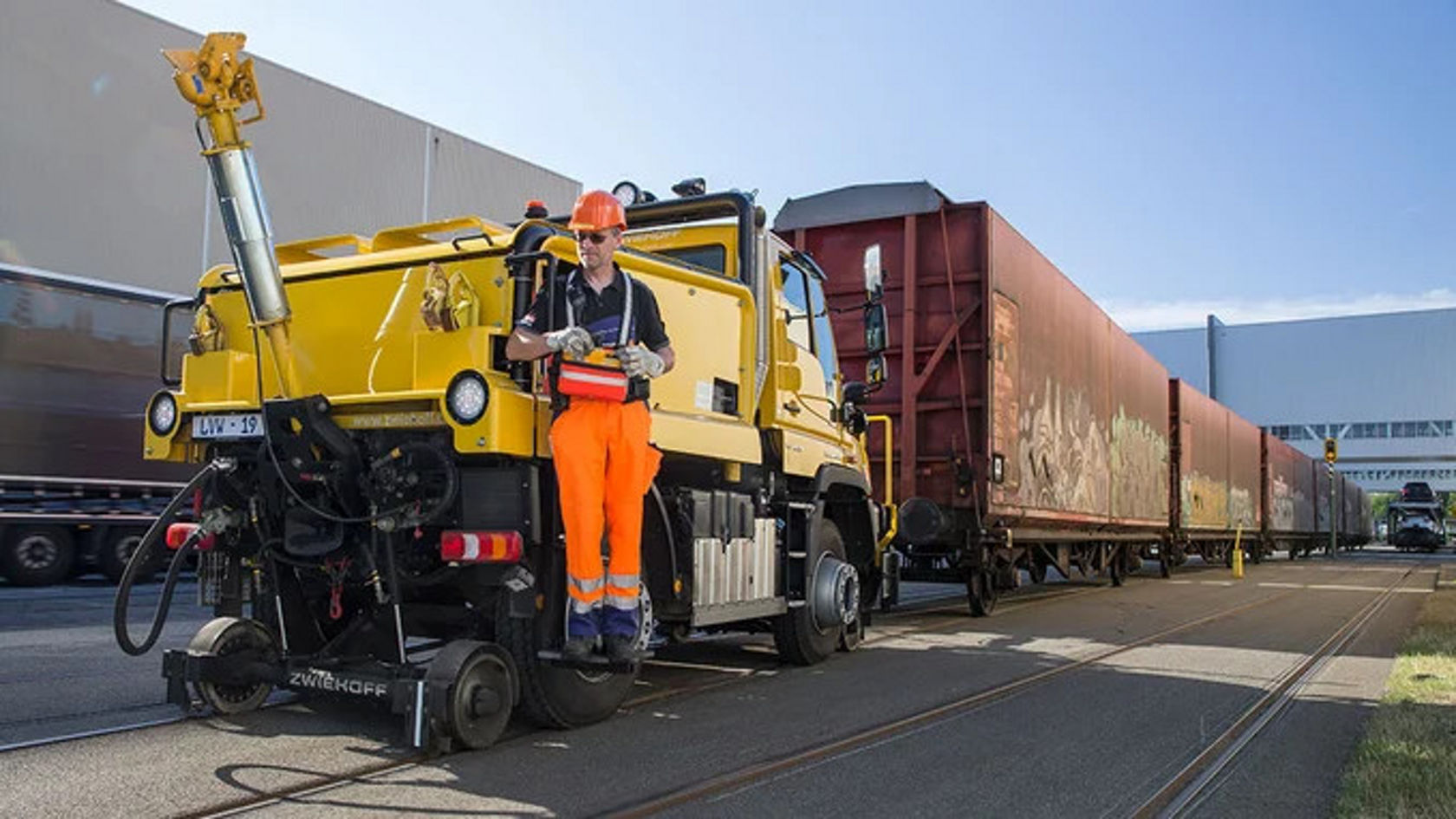 Ein gelber Unimog von Mercedes-Benz ist auf Schienen im Einsatz, während ein Arbeiter in orangefarbener Arbeitskleidung darauf steht und die Maschine bedient.