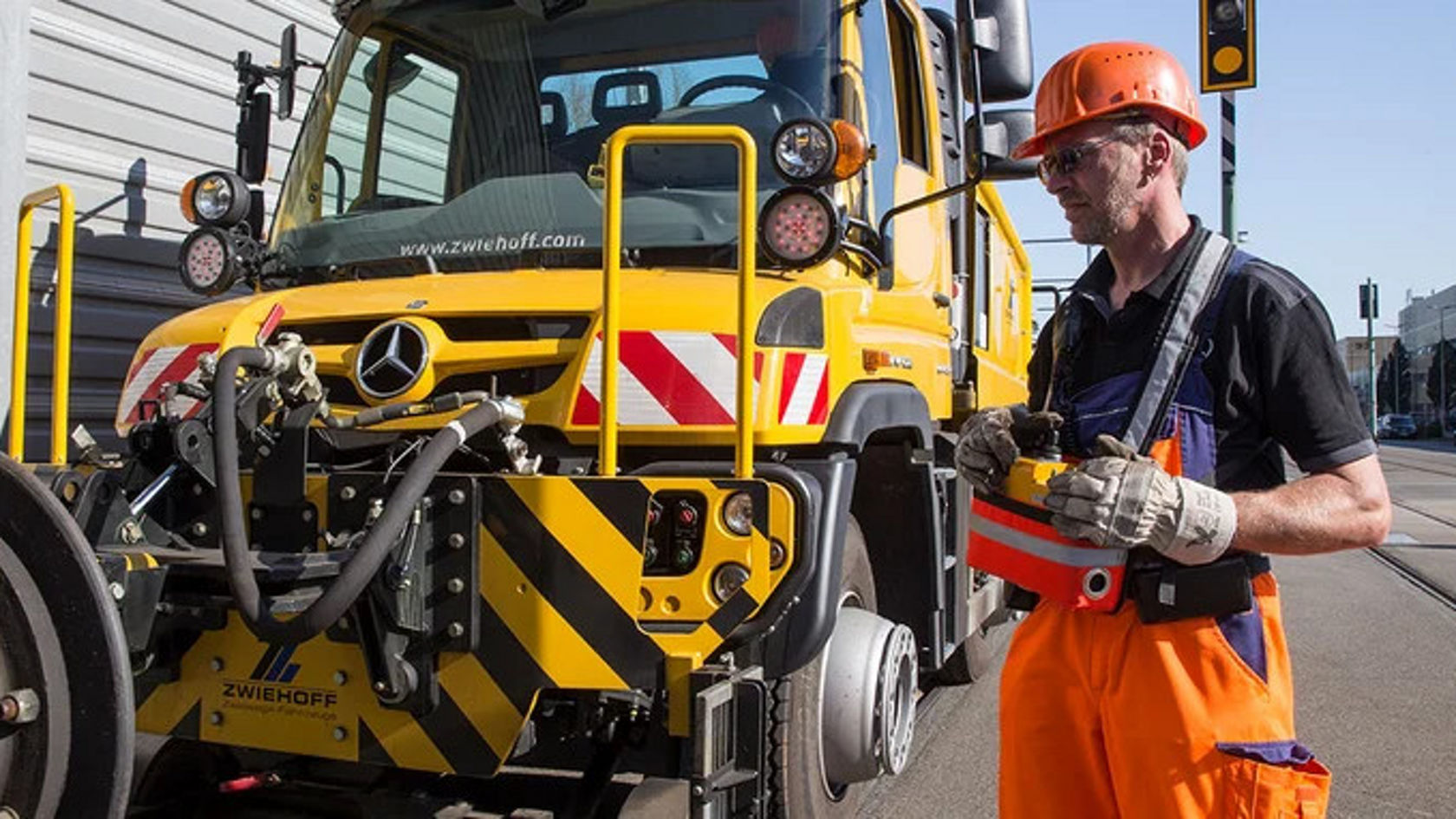 Ein Arbeiter in orangefarbener Arbeitskleidung steht neben einem gelben Mercedes-Benz Unimog, während er ein Werkzeug hält und sich auf die Ausrüstung konzentriert.