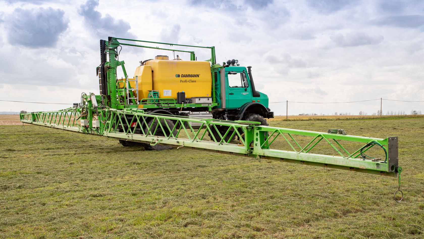 Ein grüner Sprühtraktor mit einem großen gelben Tank auf dem Anhänger steht auf einem Feld. Die Sprühvorrichtung ist ausgeklappt und zeigt eine landwirtschaftliche Anwendung.
