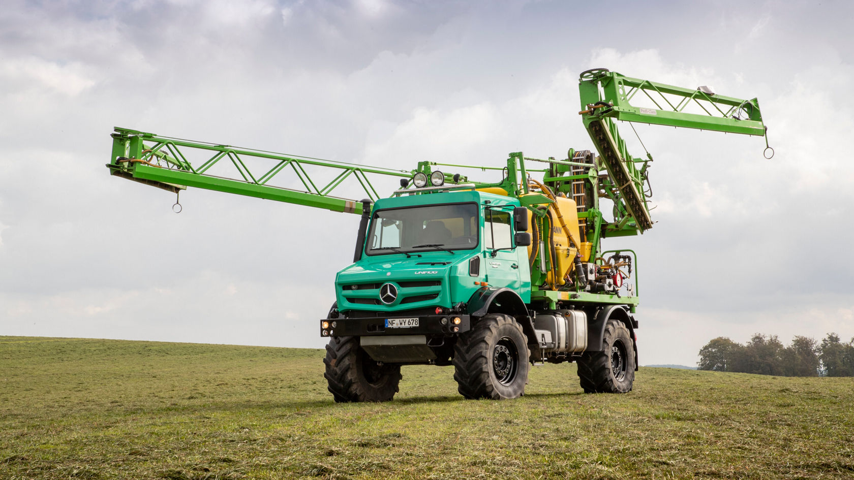 Ein grünes, geländegängiges Fahrzeug von Mercedes-Benz mit ausgefahrenem Kranarm steht auf einer Wiese, bereit für landwirtschaftliche Arbeiten.