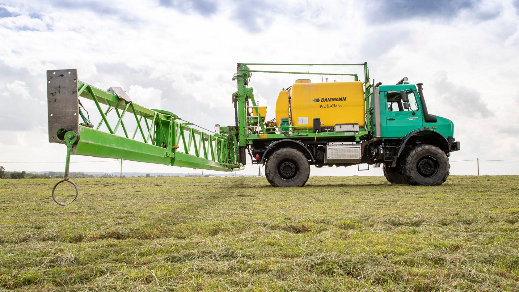 Ein grüner Sprühtraktor mit einem langen, grünen Ausleger und einem gelben Wassertank steht auf einer Wiese. Das Fahrzeug ist für landwirtschaftliche Anwendungen konzipiert.