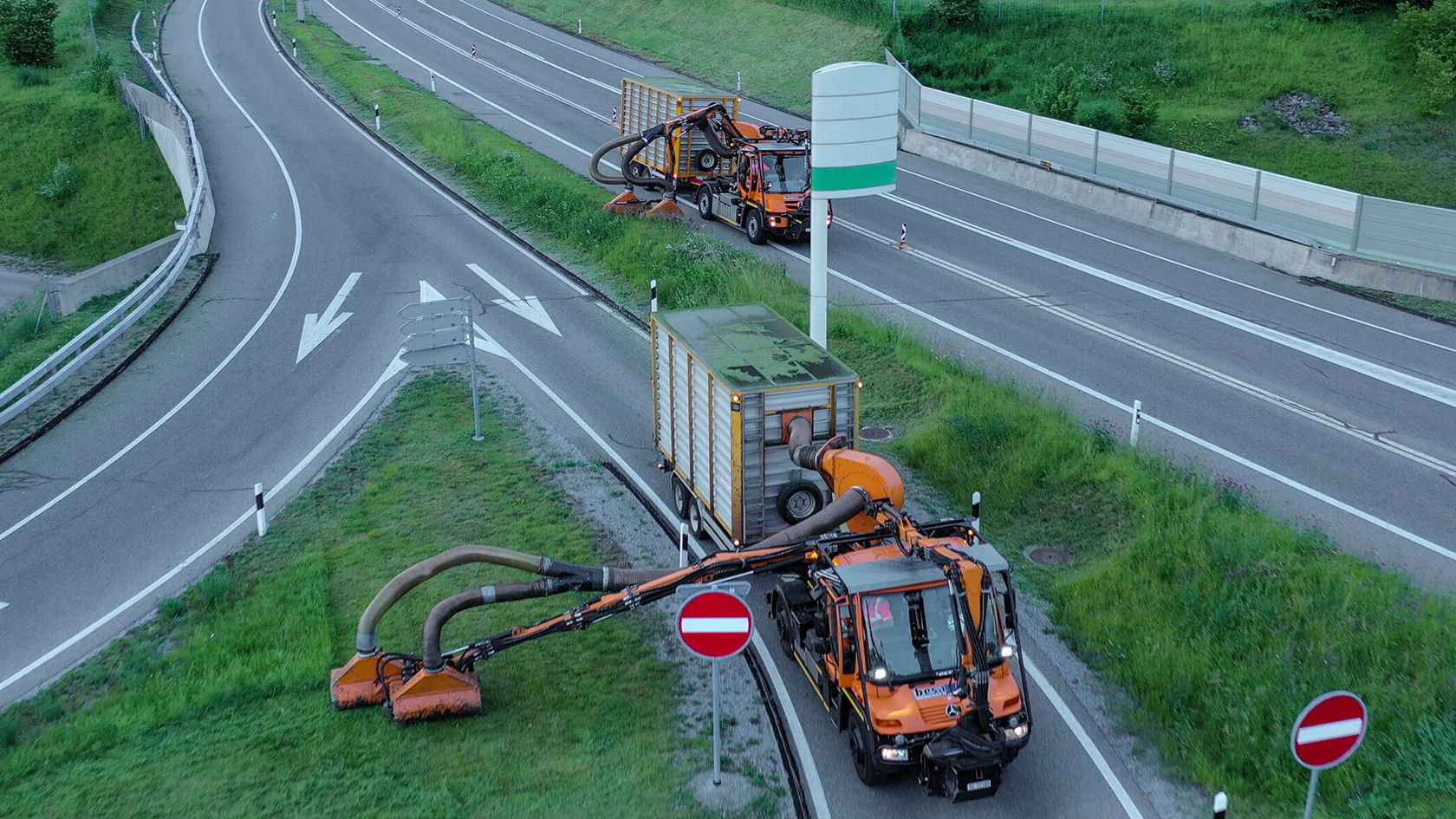 Zwei orangefarbene Maschinen, ausgestattet mit großen Schläuchen, arbeiten auf einer Straßenabzweigung zur Straßenreinigung. Im Hintergrund sind Verkehrsschilder zu sehen.