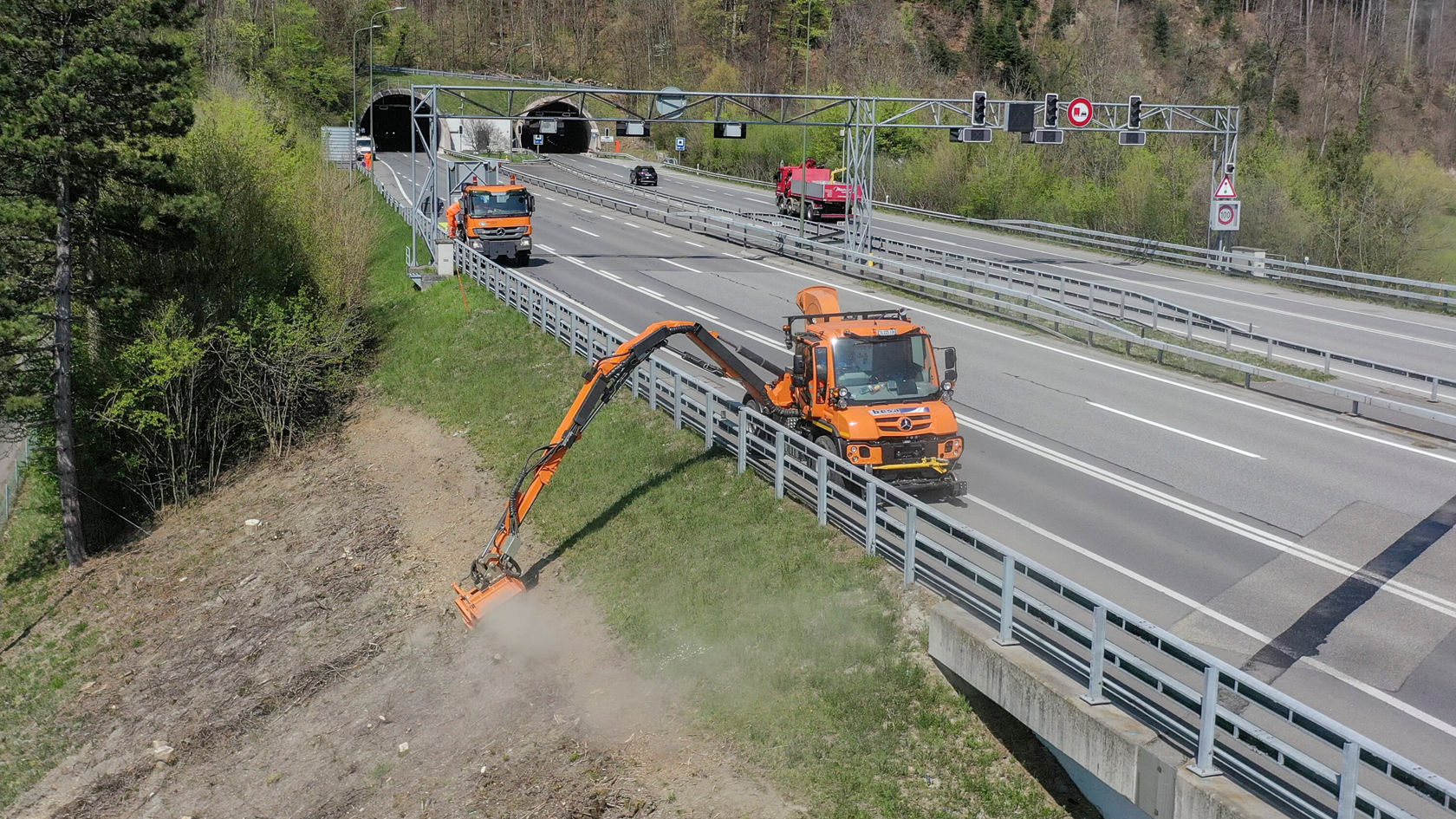 Zwei orangefarbene Mercedes-Benz Fahrzeuge arbeiten an einer Baustelle neben einer Straße, während ein Rasenmäherarm über das Gras schneidet. Im Hintergrund ist ein Tunnel und Verkehrsschilder sichtbar.