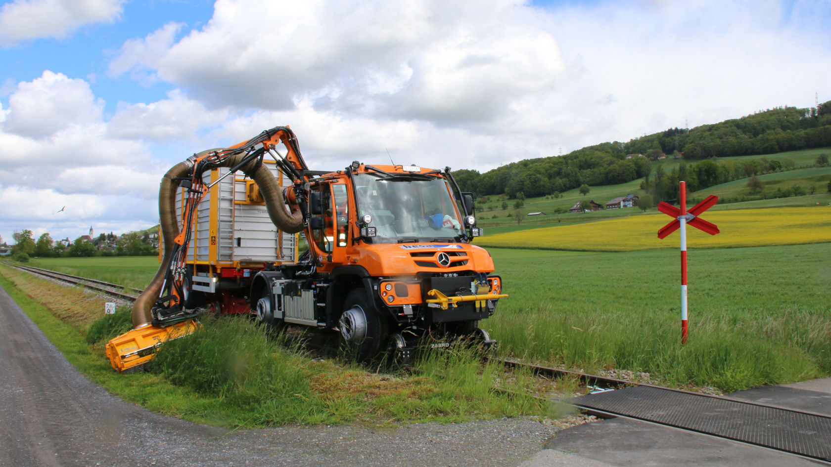 Ein orangefarbener Mercedes-Benz Arbeitsfahrzeug steht neben den Bahngleisen, ausgestattet mit einem speziellen Reinigungssystem zur Pflege des Gleisbereichs, umgeben von grünen Feldern.