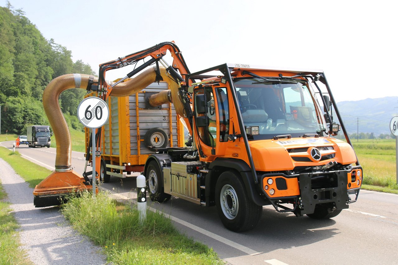 Ein orangefarbener Mercedes-Benz Einsatzfahrzeug mit einem großen Saugrohr auf einer Landstraße, das winzige Rückstände oder Abfälle aufnimmt, während ein Verkehrsschild mit der Geschwindigkeit 60 im Vordergrund sichtbar ist.