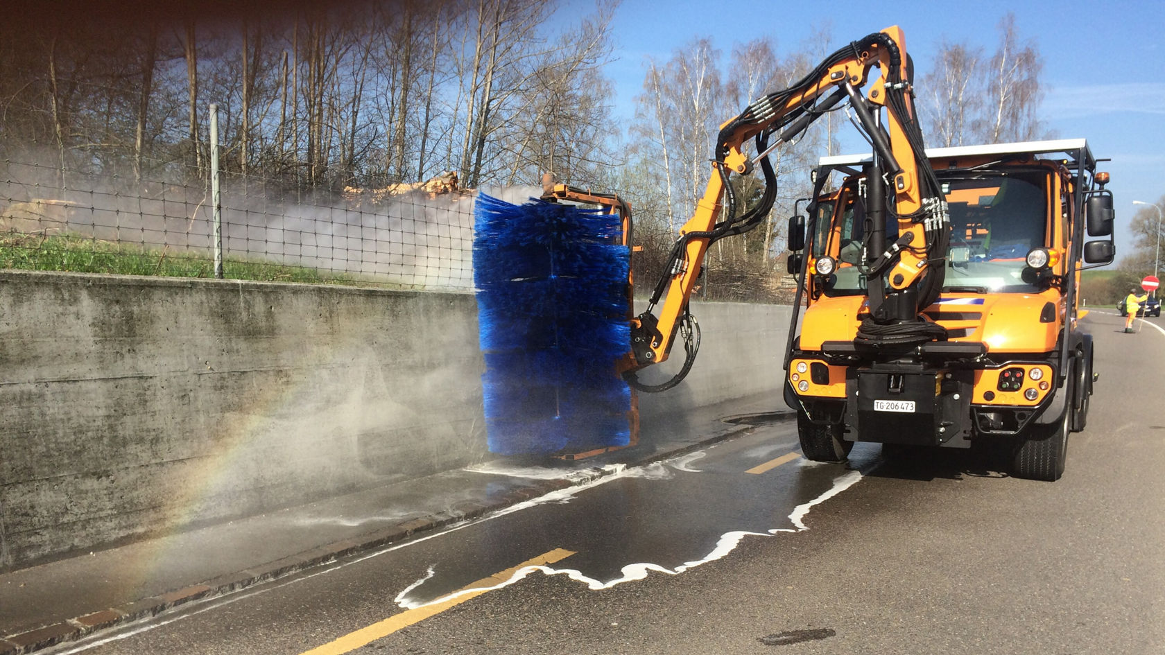 Ein Spezialfahrzeug von Mercedes-Benz reinigt den Straßenrand mit einem Wasserstrahl und einer großen rotierenden Bürste, während Dampf aufsteigt und ein klarer Himmel sichtbar ist.