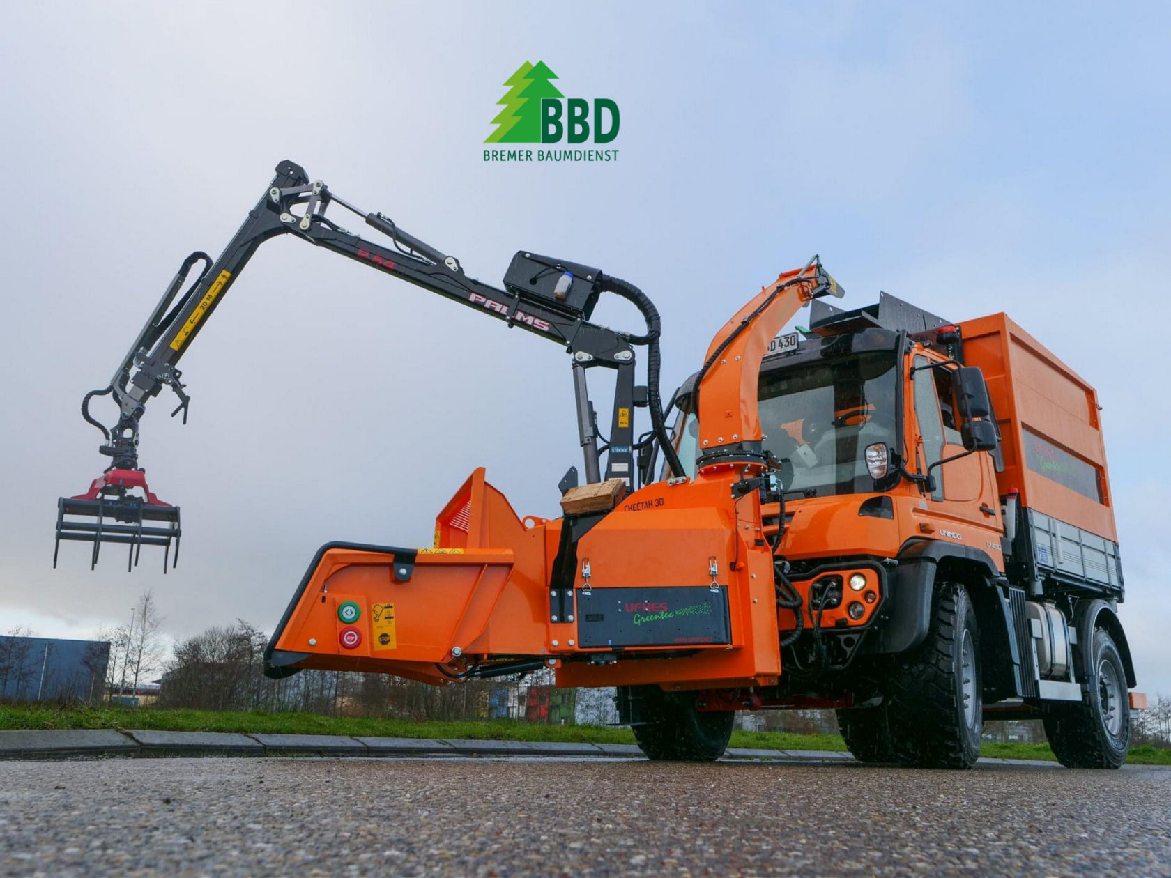 Un véhicule orange de Bremer Baumschnittdienst, équipé d'une broyeuse à bois, se trouve dans la neige. Le véhicule porte le logo et les coordonnées de l'entreprise sur le côté.