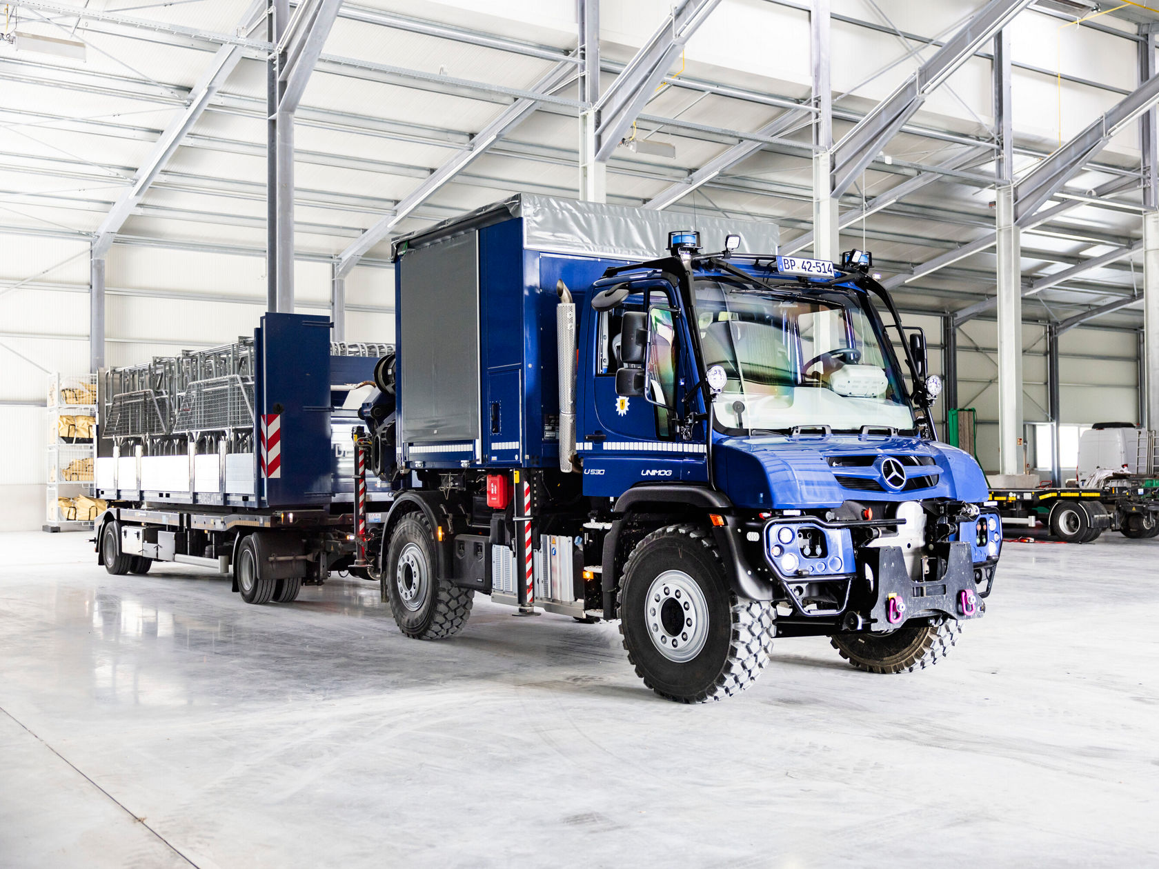Ein blauer Mercedes-Benz Unimog mit Anhänger steht in einer Lagerhalle.