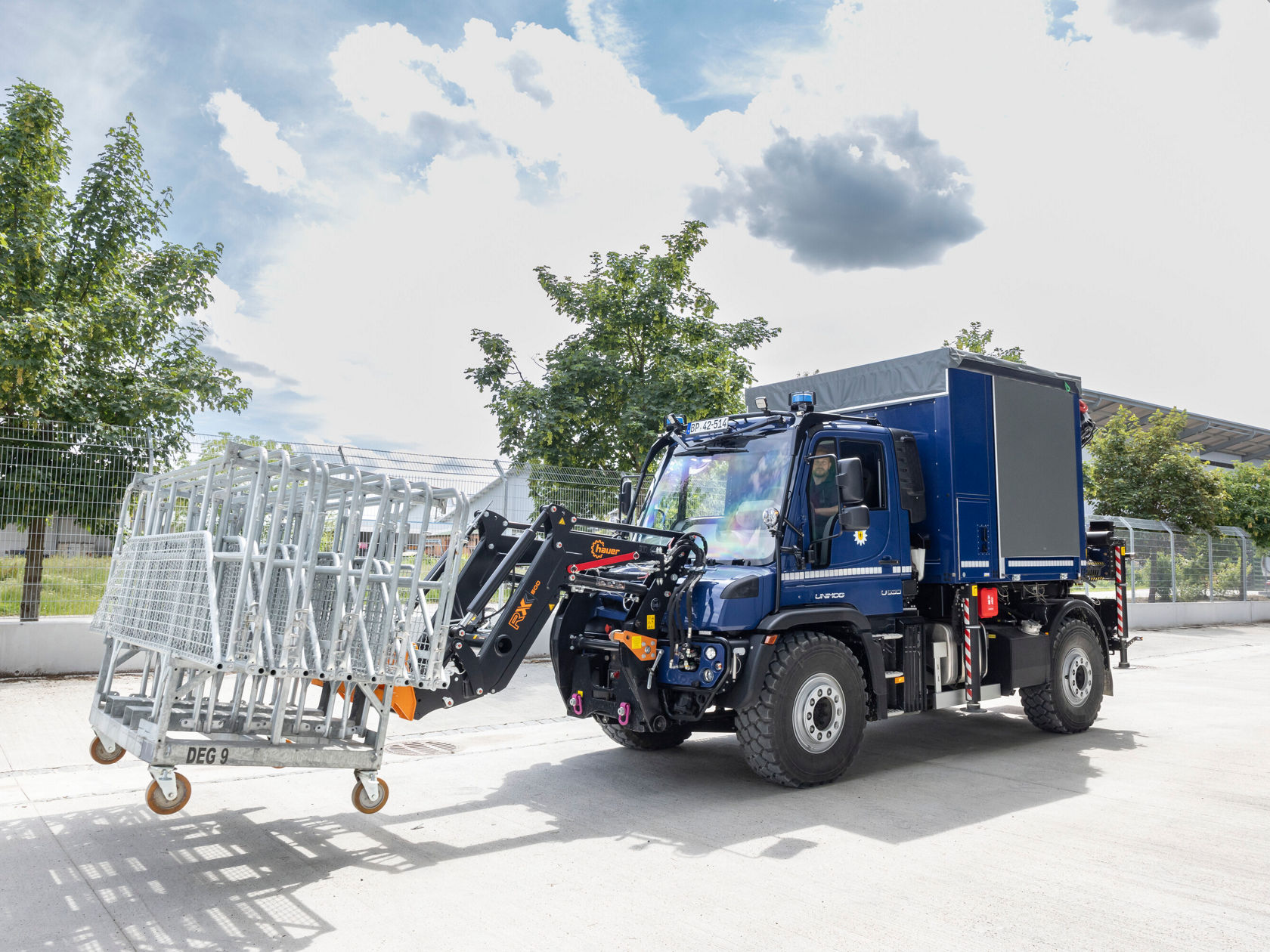 Ein Mercedes-Benz Unimog hebt mit einem Greifarm ein Gestell aus Metallgestelle.
