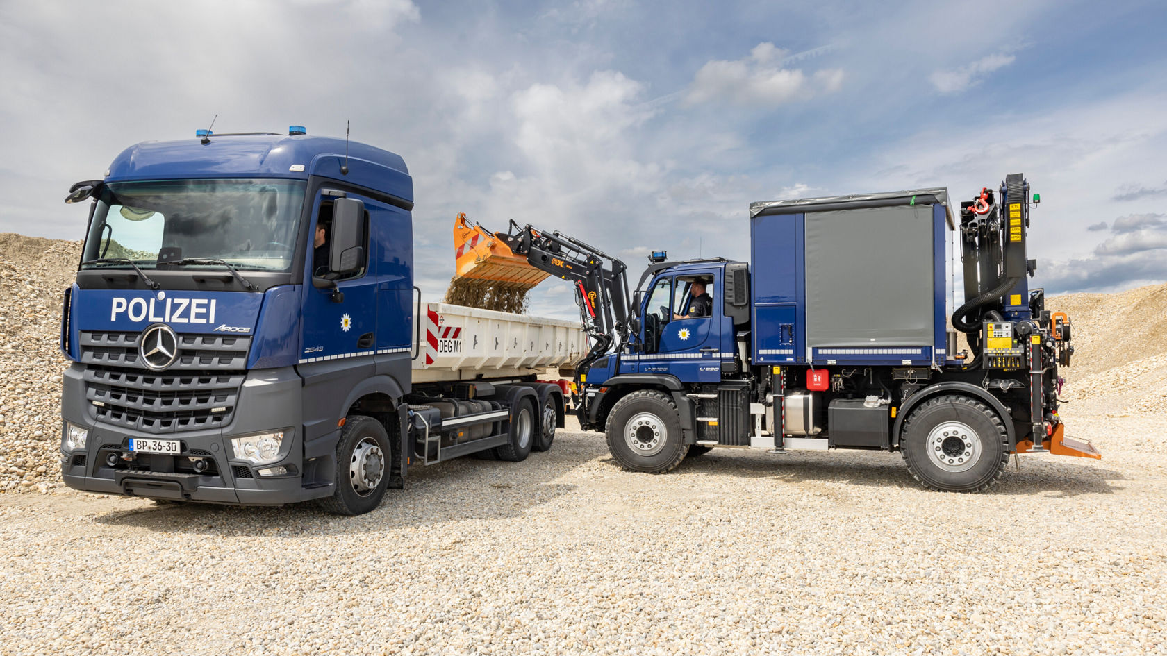 Un camion Mercedes‑Benz portant l'inscription « POLICE » est garé à côté d'un Unimog équipé d'une plate-forme de chargement.