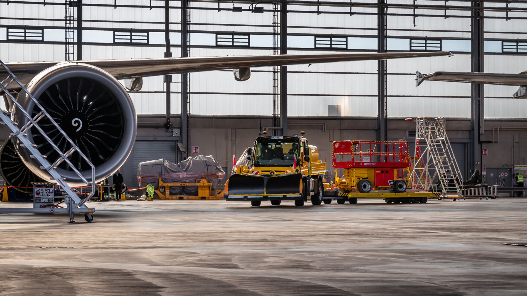 Eine große Flugzeugturbine befindet sich im Vordergrund, während ein gelbes Servicefahrzeug und eine Hebebühne in einer Flughafeninstandhaltungswerkstatt zu sehen sind.