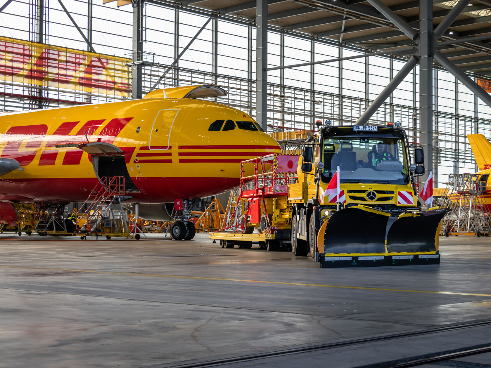 Ein gelbes Mercedes-Benz Fahrzeug mit Schneeschaufel steht in einem Hangar, umgeben von DHL-Frachtflugzeugen. Die Umgebung zeigt eine helle und gut strukturierte Wartungsanlage.