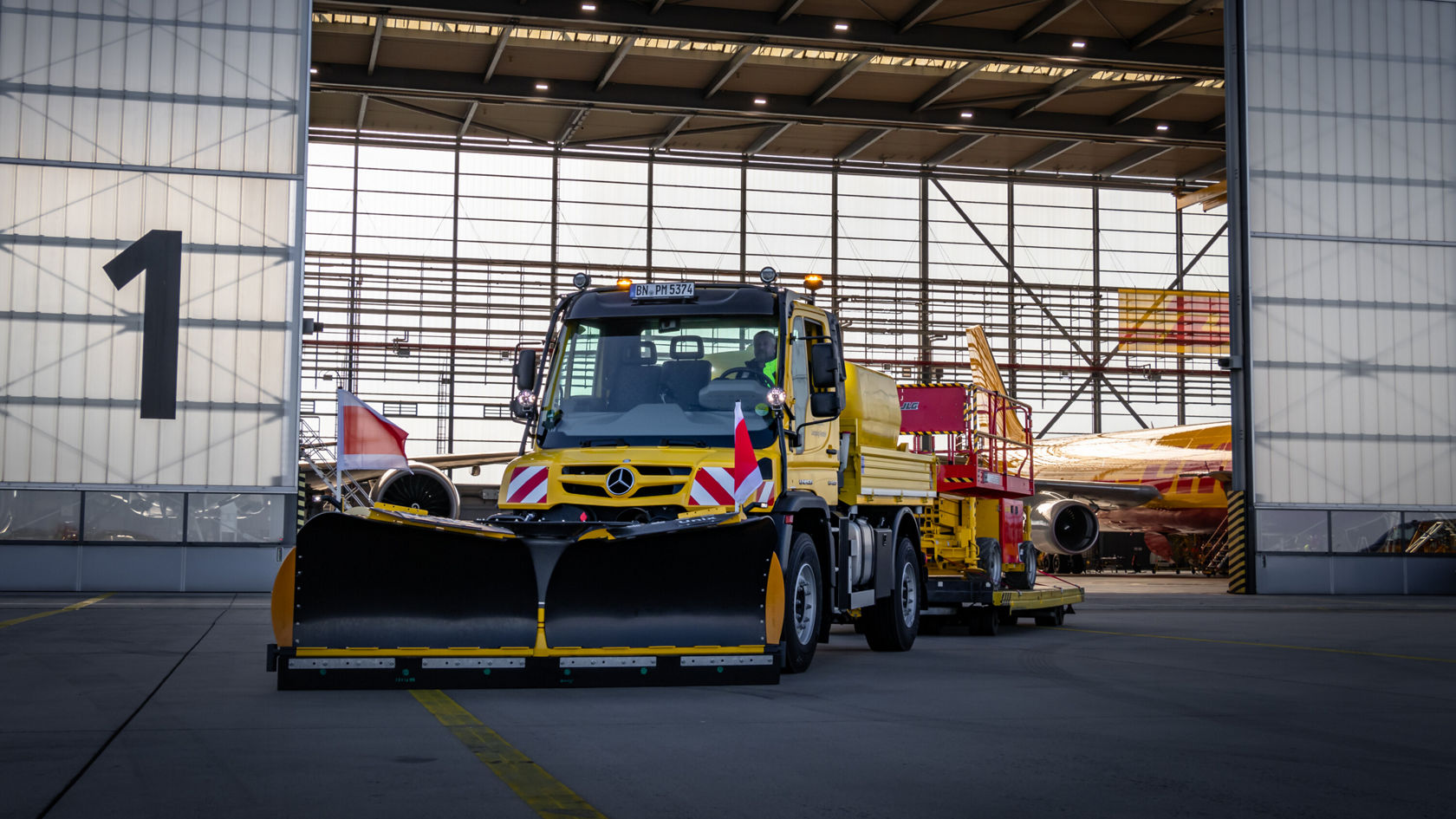 Ein gelber Mercedes-Benz-Lkw steht mit einem Schneepflug und einem Anhänger vor einem Hangar, bereit für Einsätze am Flughafen.