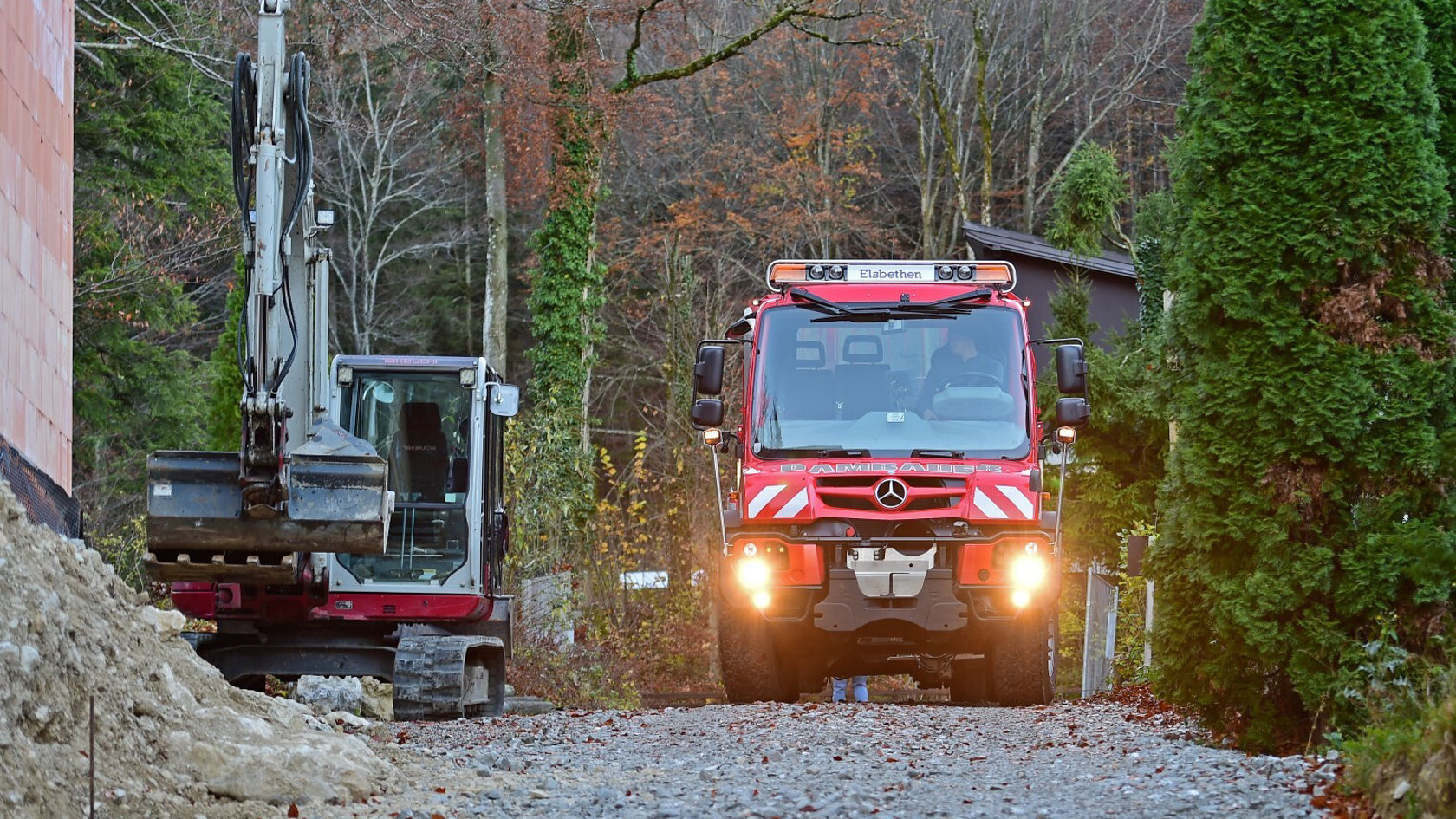 Ein roter Mercedes-Benz Feuerwehrwagen fährt auf einem schotterigen Weg, während ein Bagger auf der linken Seite geparkt ist, umgeben von Bäumen und einer Baustelle im Hintergrund.