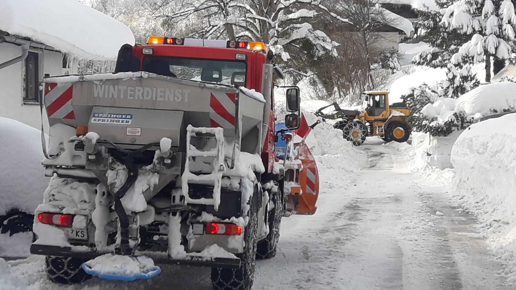 Ein Mercedes-Benz Winterdienstfahrzeug mit Schneepflug räumt eine verschneite Straße, während im Hintergrund ein Bagger Schnee beseitigt.