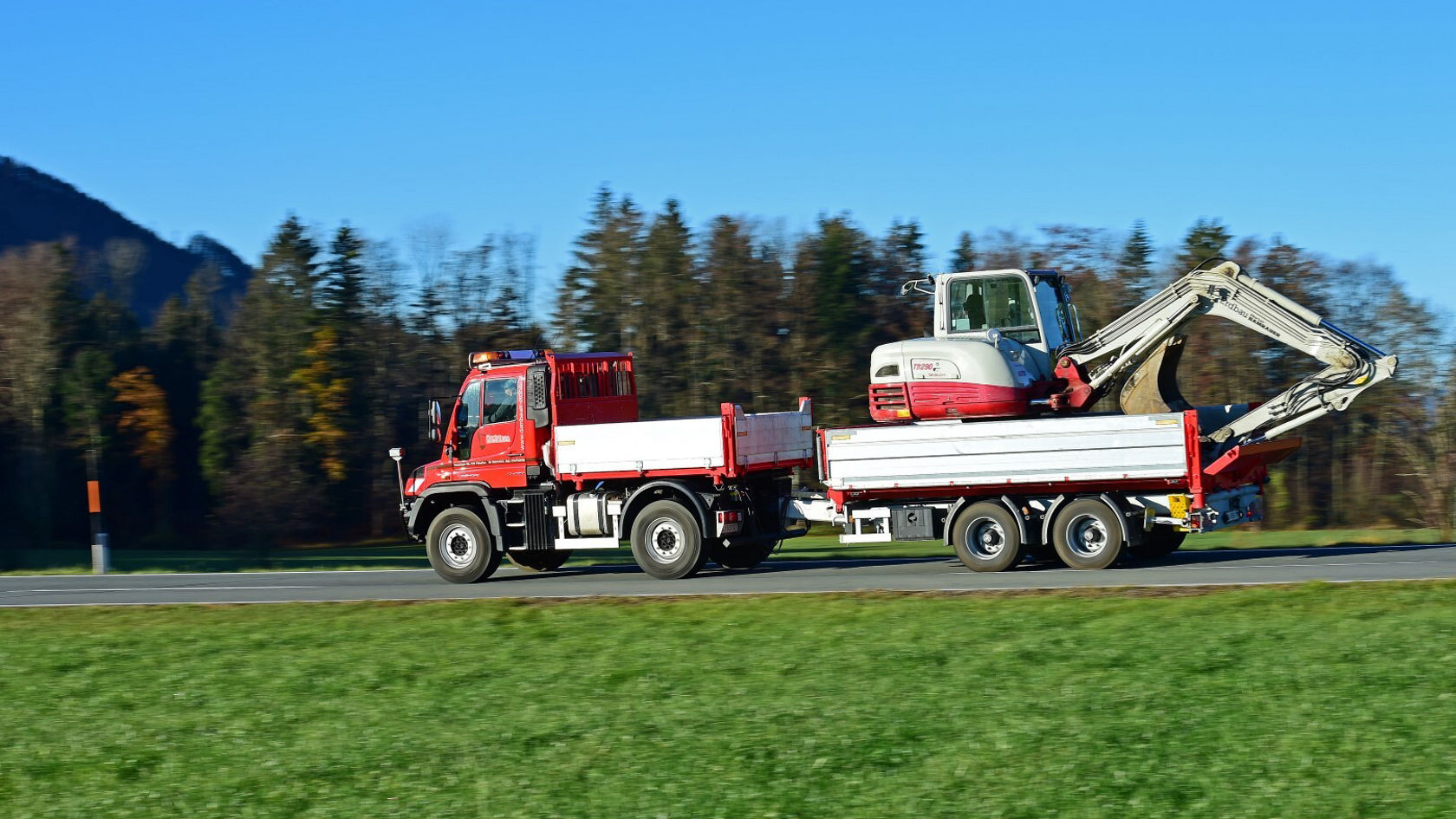 Ein rotes Nutzfahrzeug mit Anhänger transportiert einen Bagger auf einer Straße, umgeben von grüner Wiese und Bäumen im Hintergrund.