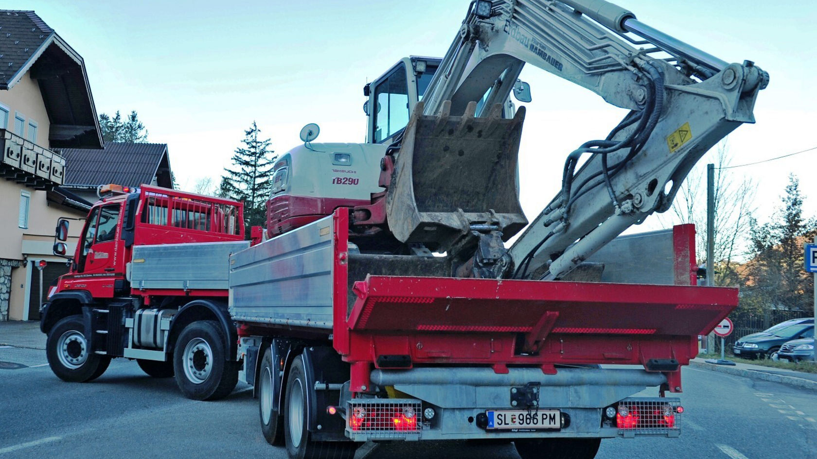 Ein roter Mercedes-Benz Lkw mit einer Baumaschine auf der Ladefläche steht auf einer Straße. Die Baumaschine hat einen großen Baggerarm, der in Richtung Himmel zeigt.