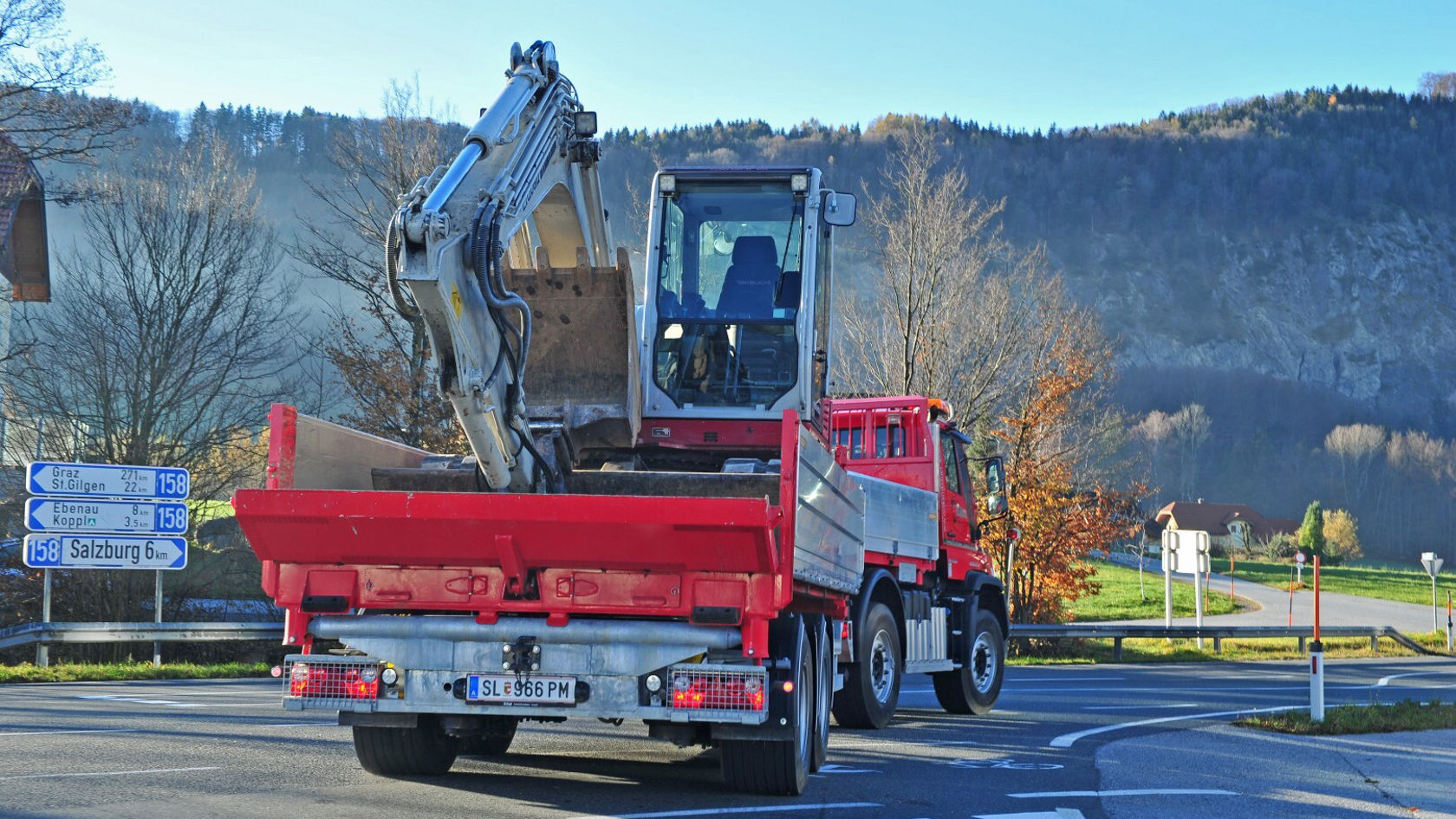 Ein roter Mercedes-Benz Lkw steht an einer Kurve, mit einem Bagger auf der Ladefläche, bereit für den Einsatz auf einer Baustelle.