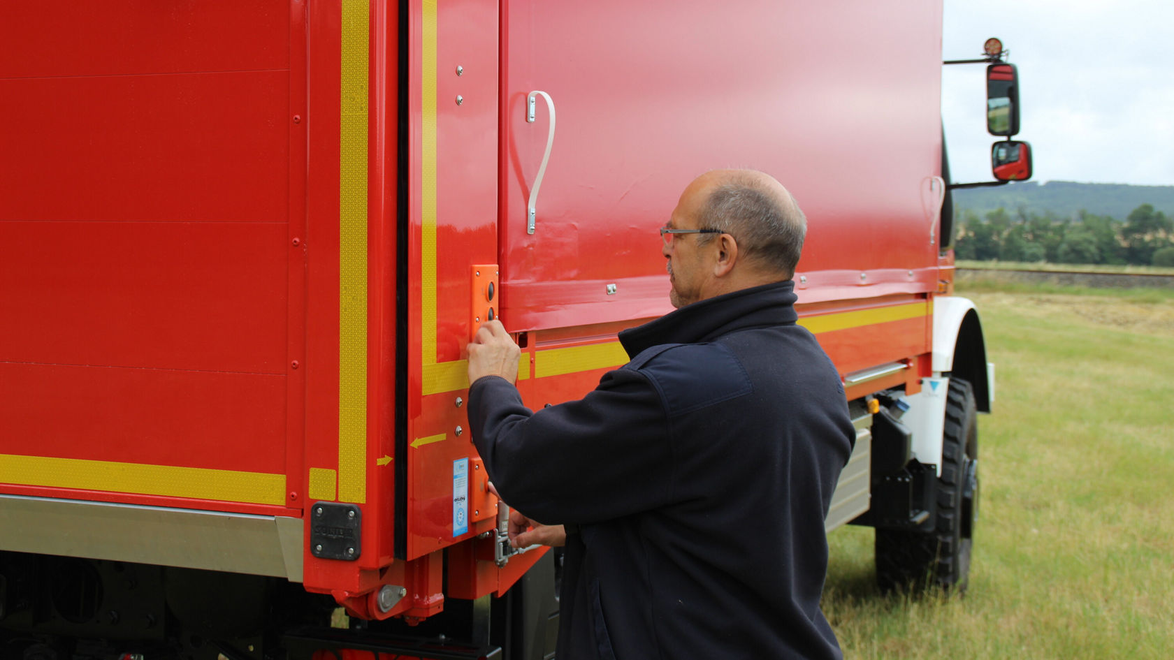 Ein Mann in einer schwarzen Jacke schließt eine Tür an einem roten Mercedes-Benz Lkw auf einem offenen Feld.