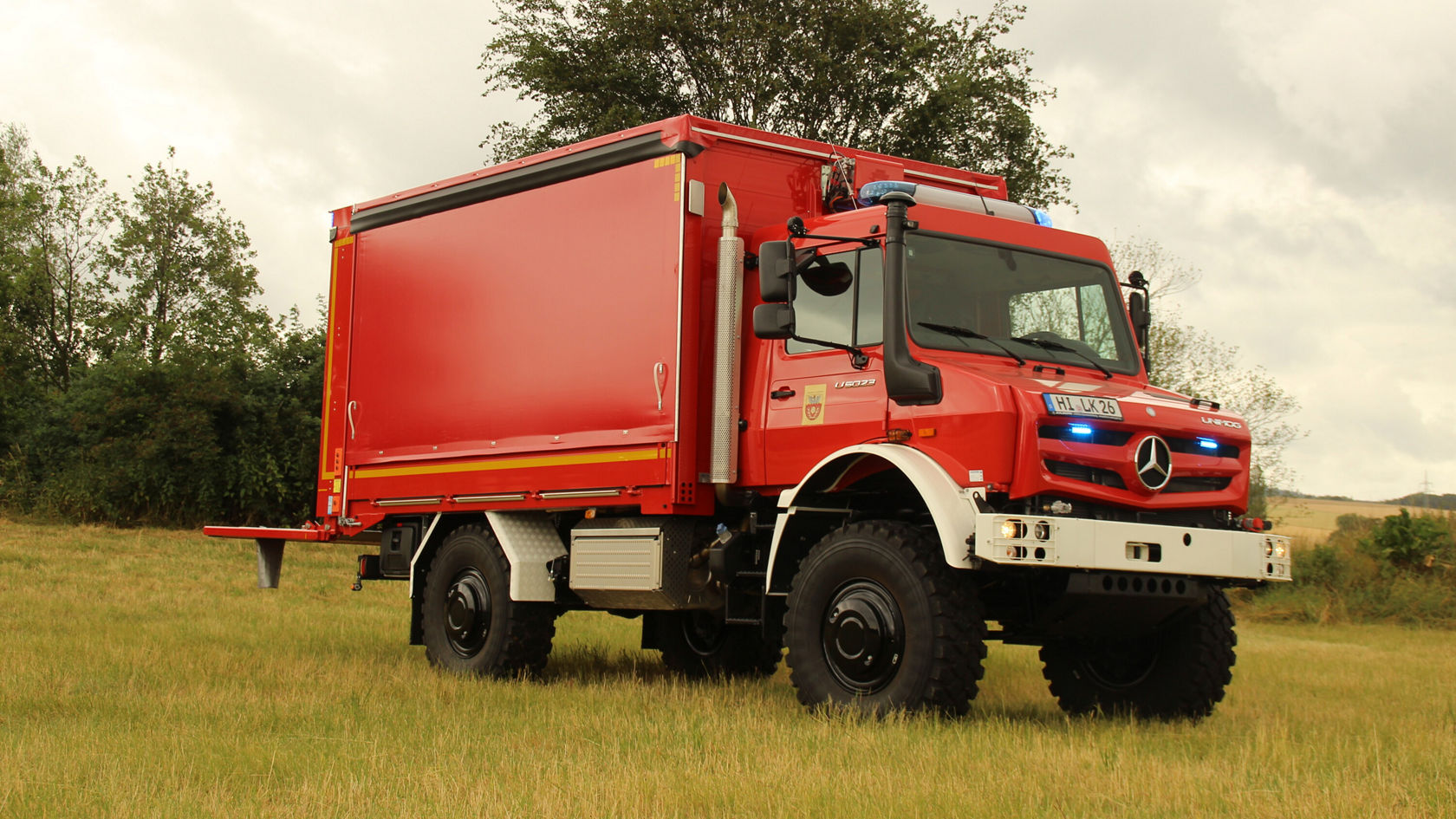 Roter Mercedes-Benz Unimog mit Lkw-Kabine steht auf einer Wiese, ausgestattet mit Blaulicht und einer robusten Ladefläche, ideal für Einsatzfahrten oder Transport.