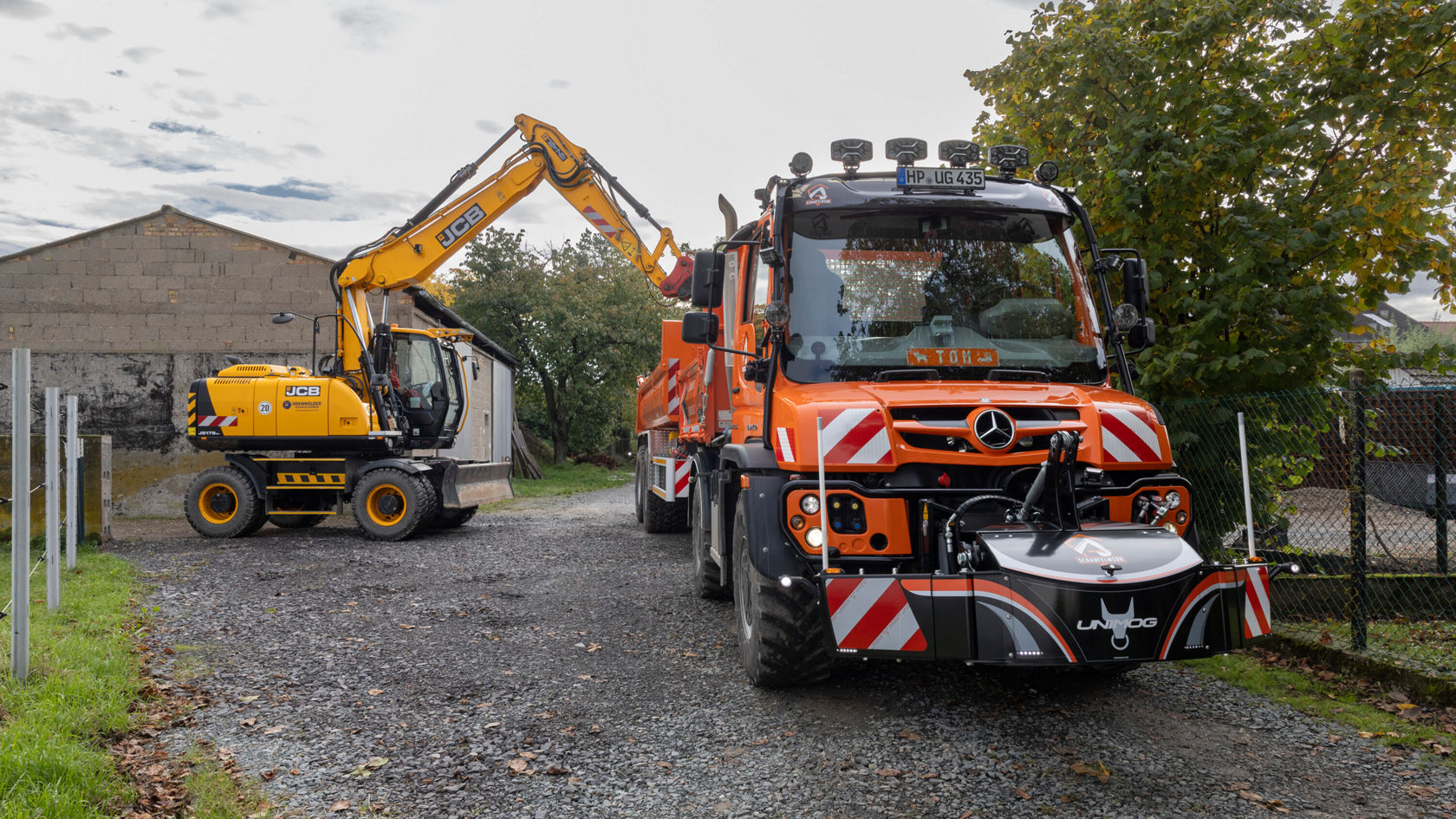 Un camion Mercedes‑Benz orange se trouve sur une route de gravier à côté d'une pelleteuse jaune équipée d'un godet, orientée vers une extension voisine pour effectuer des travaux de construction.