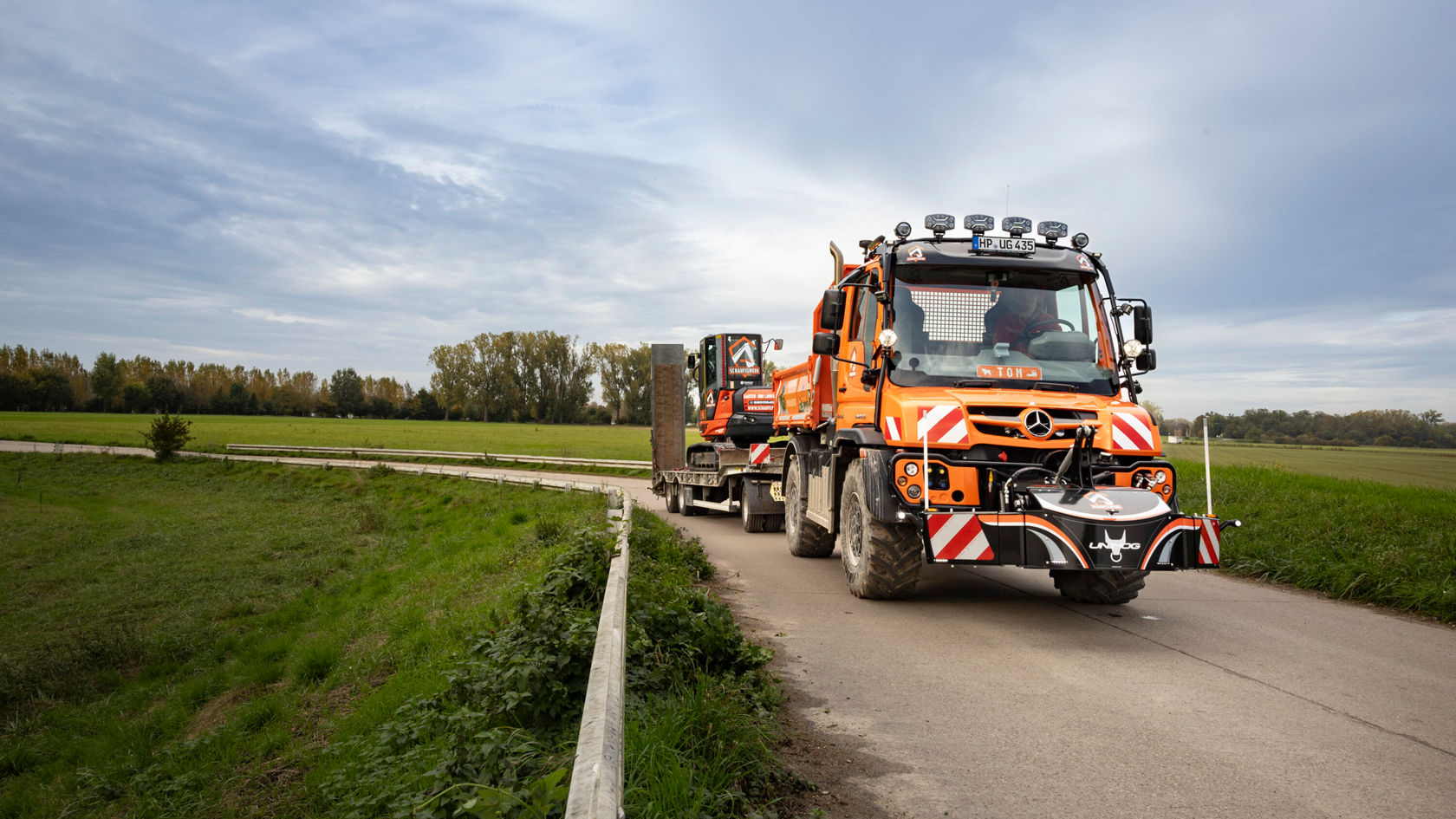 Un tout-terrain Mercedes‑Benz orange roule sur une route le long d'un champ et tracte une remorque chargée d'un équipement de travail.