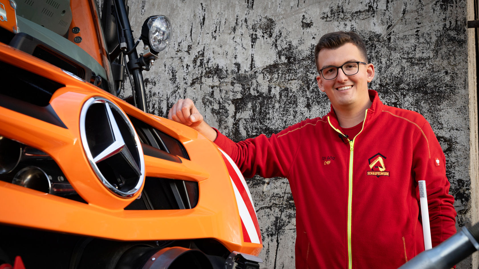Un jeune homme portant des lunettes se tient, souriant, à côté d'un camion orange Mercedes‑Benz. Il porte une veste rouge avec un logo.