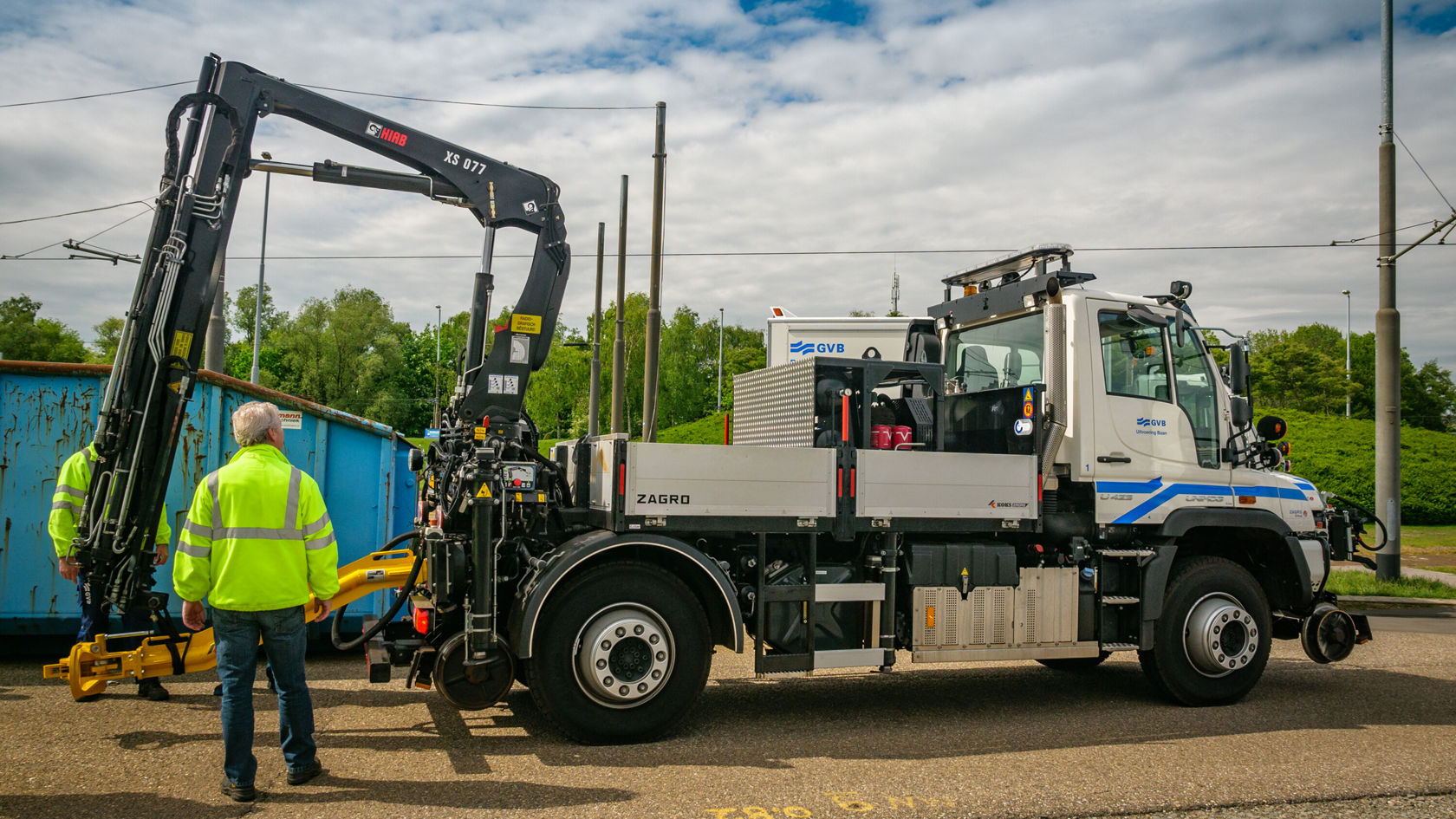 Ein Mercedes-Benz Unimog mit einem Kranaufbau wird von zwei Personen in leuchtenden Sicherheitswesten inspiziert. Im Hintergrund ist ein Container sichtbar.