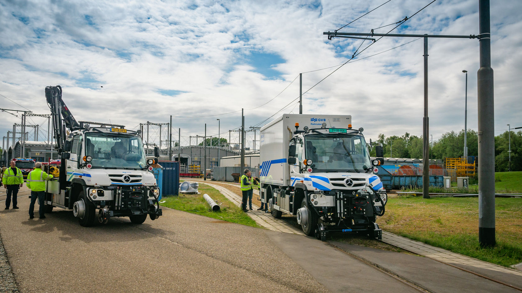 Zwei Mercedes-Benz Unimog Lastwagen stehen auf einem Gelände, während mehrere Personen in Arbeitskleidung in der Nähe stehen, möglicherweise bei einer Inspektion oder Wartung.