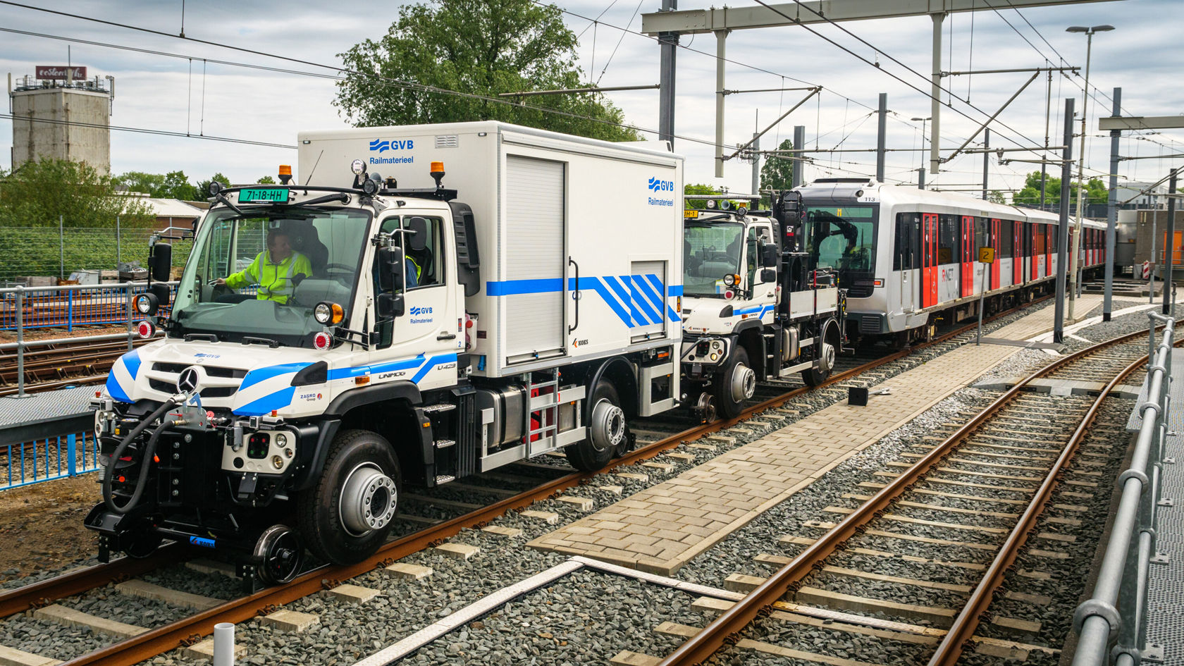 Ein Mercedes-Benz Unimog mit einem Einsatzfahrzeug steht zwischen den Schienen, während ein Stadtzug in der Nähe vorbeifährt.